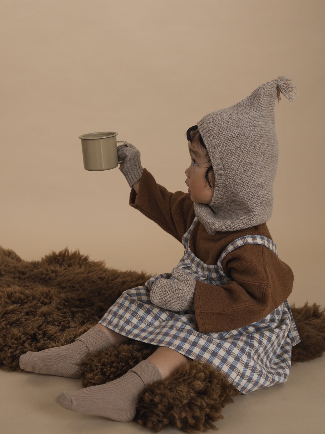 toddler in oatmeal mittens with matching balaclava, brown jumper and gingham skirt, holding cup