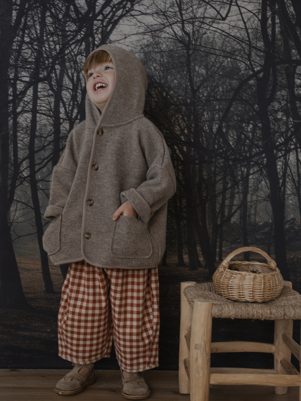 A child wearing a brownish-gray hooded coat with large front pockets, paired with red-and-white gingham pants and brown shoes, standing in front of a dark forest backdrop next to a small wooden stool with a woven basket.