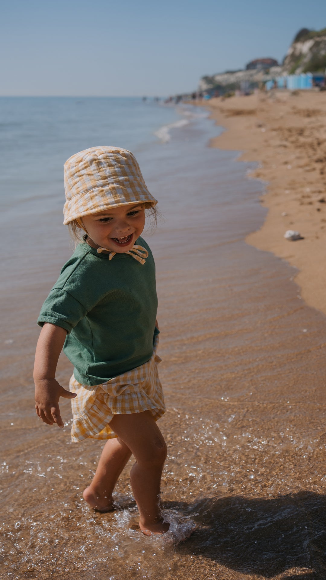 Child in a green t-shirt and hat standing on a beach with water at their feet