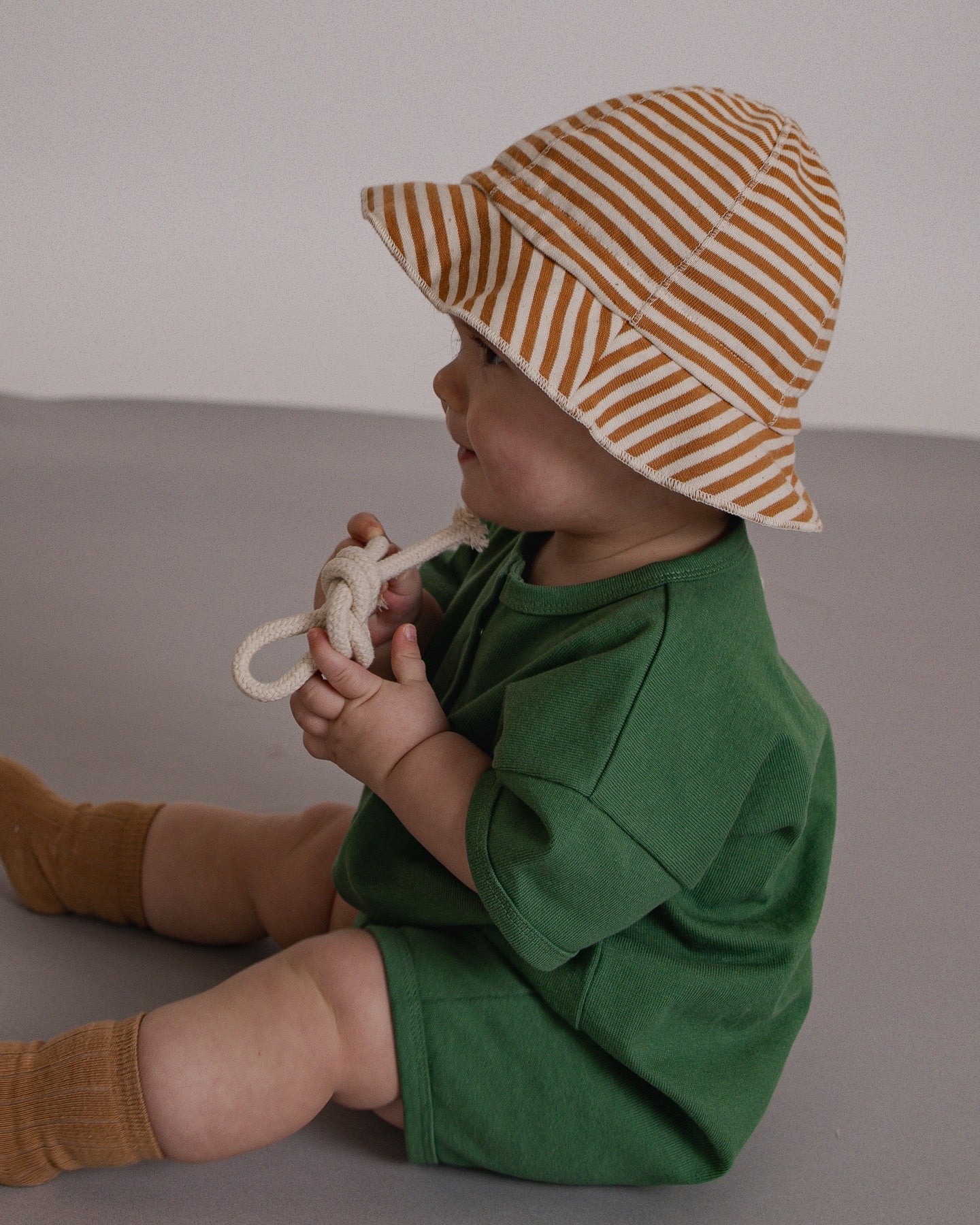 Child wearing a green outfit and striped hat sitting on a gray surface.