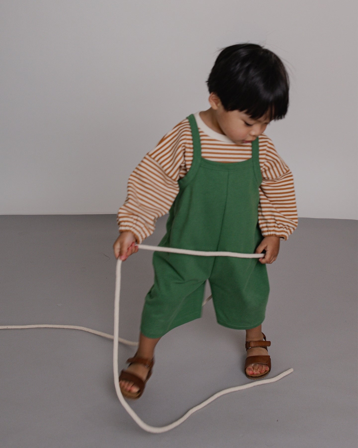 Child wearing green overalls and a striped shirt holding a long white object on a gray background