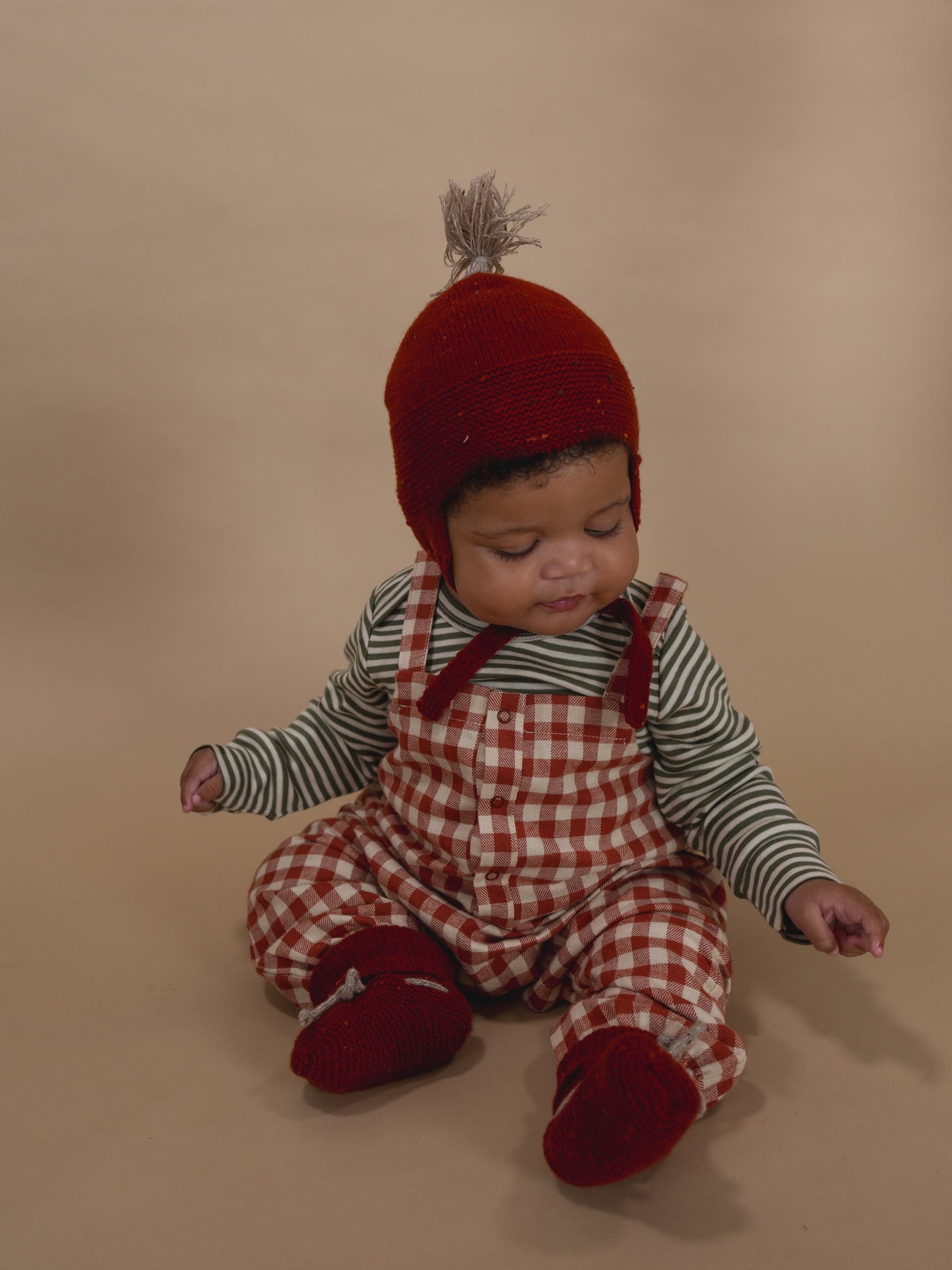 baby wearing paprika red wool booties, matching tassel hat, gingham dungarees and striped top