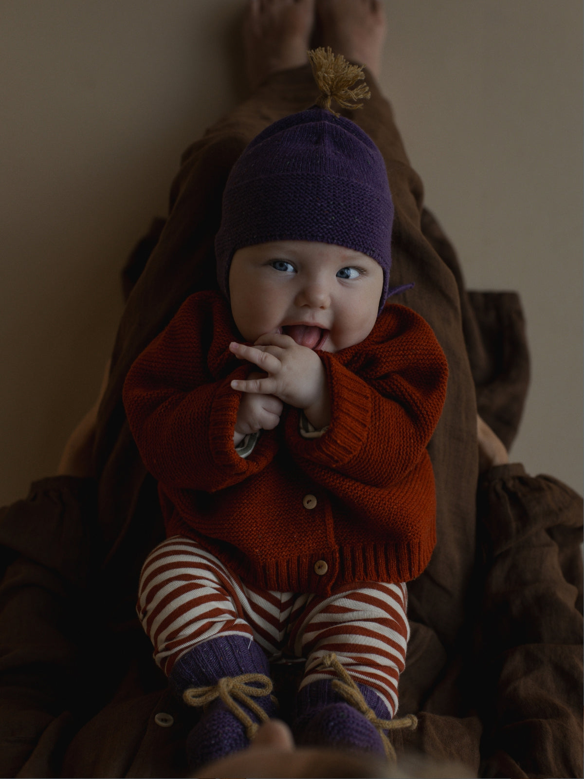 A baby wearing a purple knit bonnet with a small leaf topper, a rust-orange knit cardigan, red and white striped leggings, and purple booties, sitting on a brown blanket.