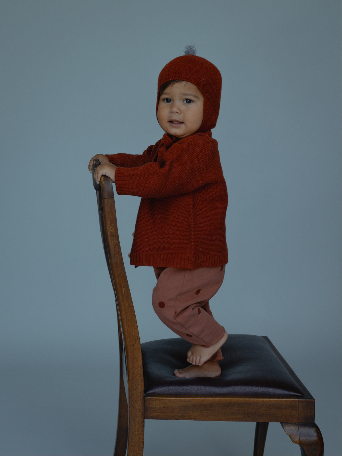 A baby standing on a wooden chair wearing a rust-orange knit hat and matching cardigan with dusty rose pants, against a solid blue-gray studio background.