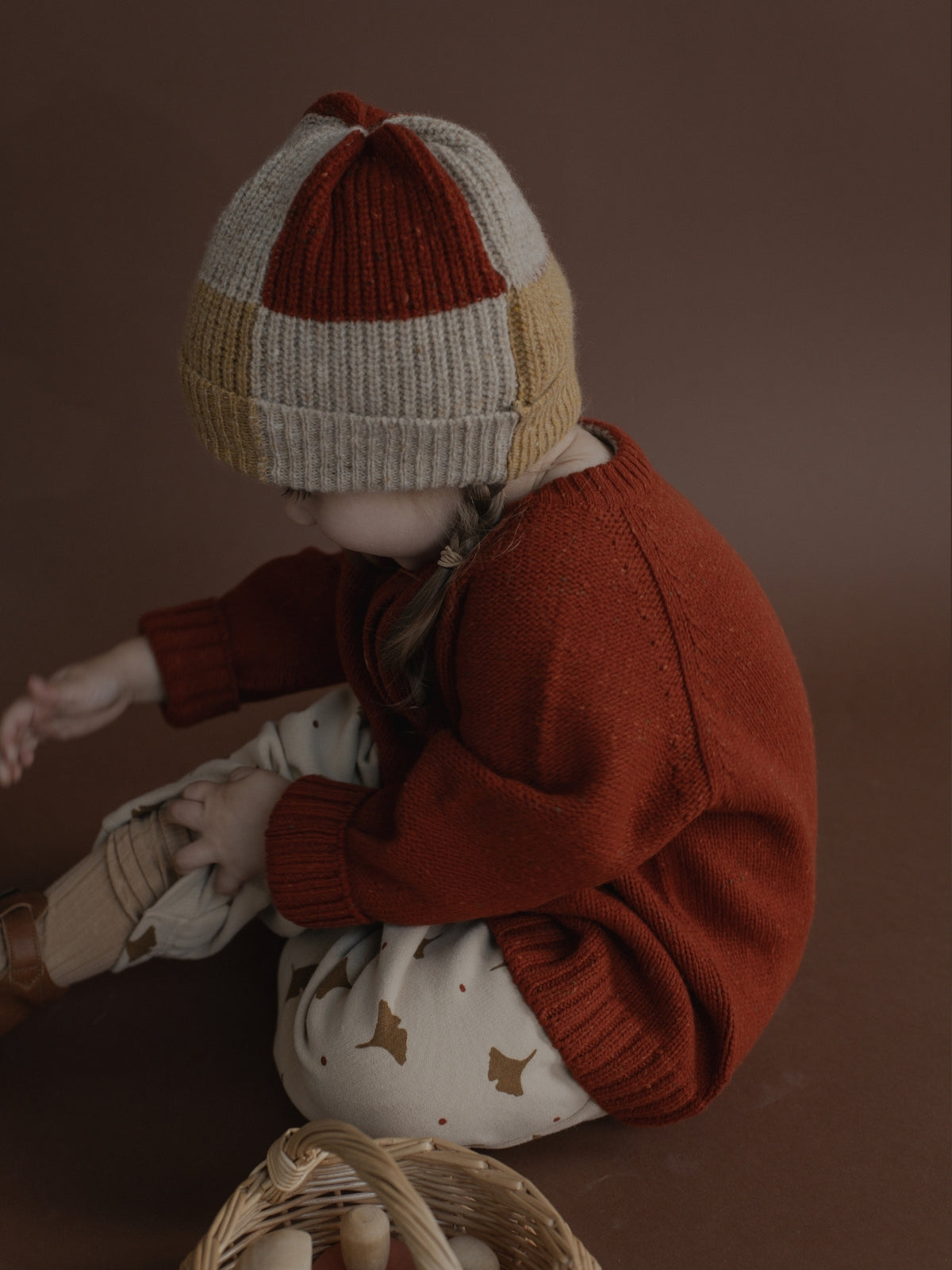 A child wearing a chunky knit beanie with rust red, cream, and tan color-blocked stripes, a rust red knit sweater, and cream pants with small brown star prints, sitting in a studio setting against a dark brown background.
