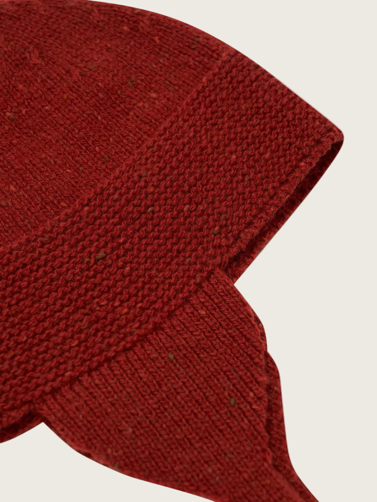 Close-up of a dark red knit beanie with a ribbed cuff and ear flaps, showing a heathered texture with tiny flecks, on a light cream background.
