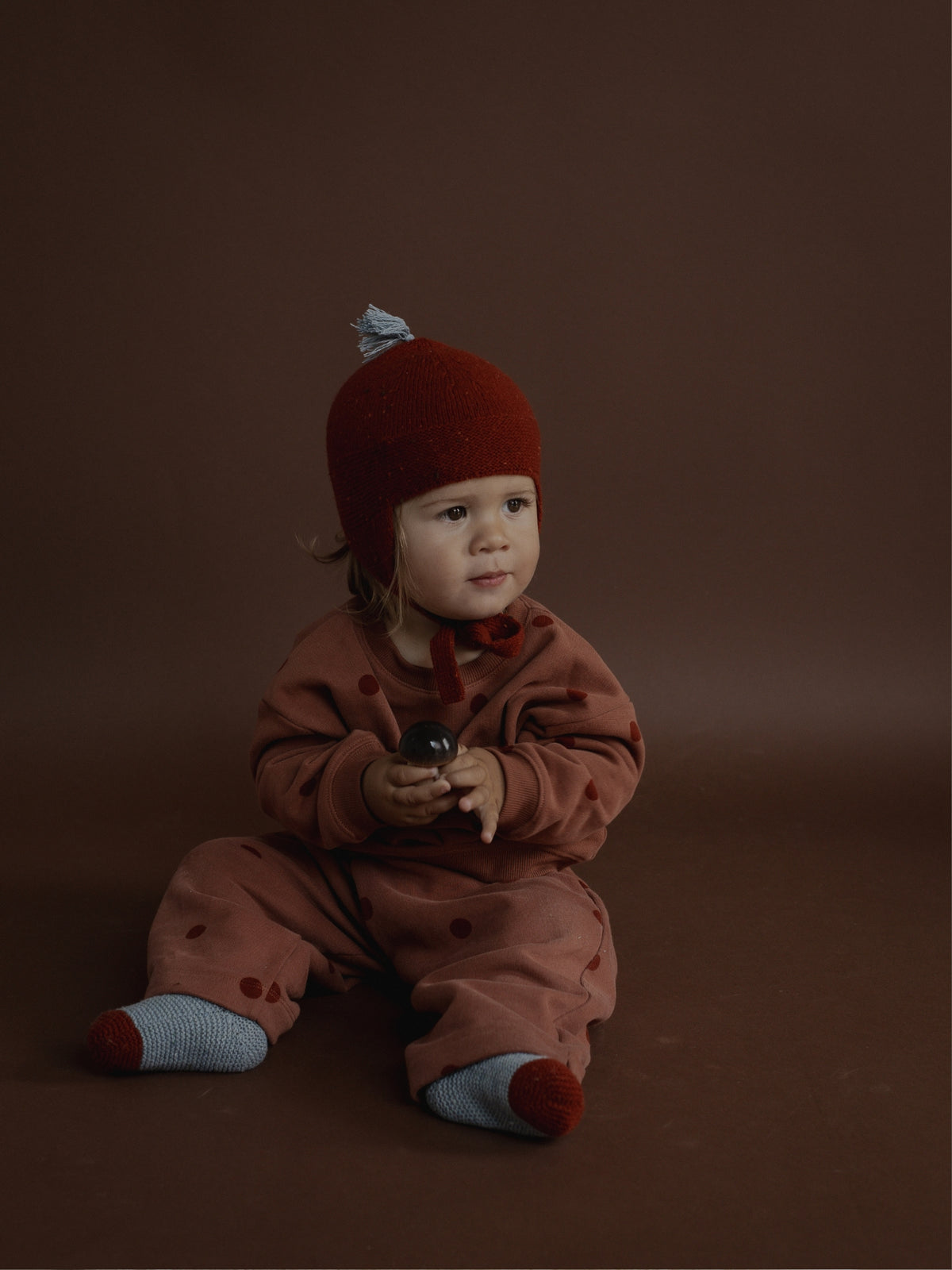 A baby sitting on a dark brown studio background, wearing a rust-brown polka-dot one-piece jumpsuit, a matching rust-brown knit hat with a small gray pom-pom, and gray socks with rust-brown toes.