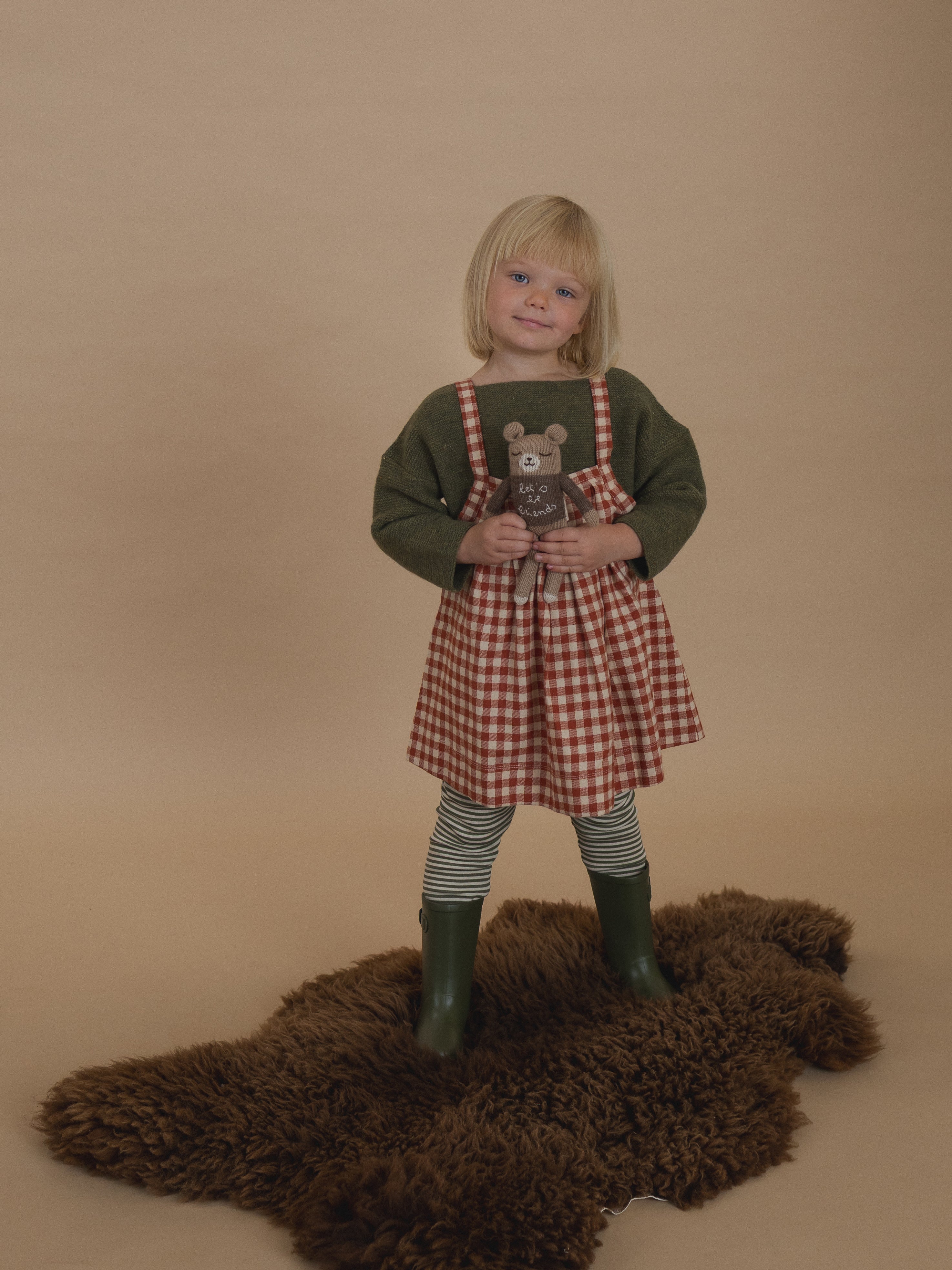 toddler in red gingham pinafore skirt with wool jumper, holding a plushie and standing on a rug