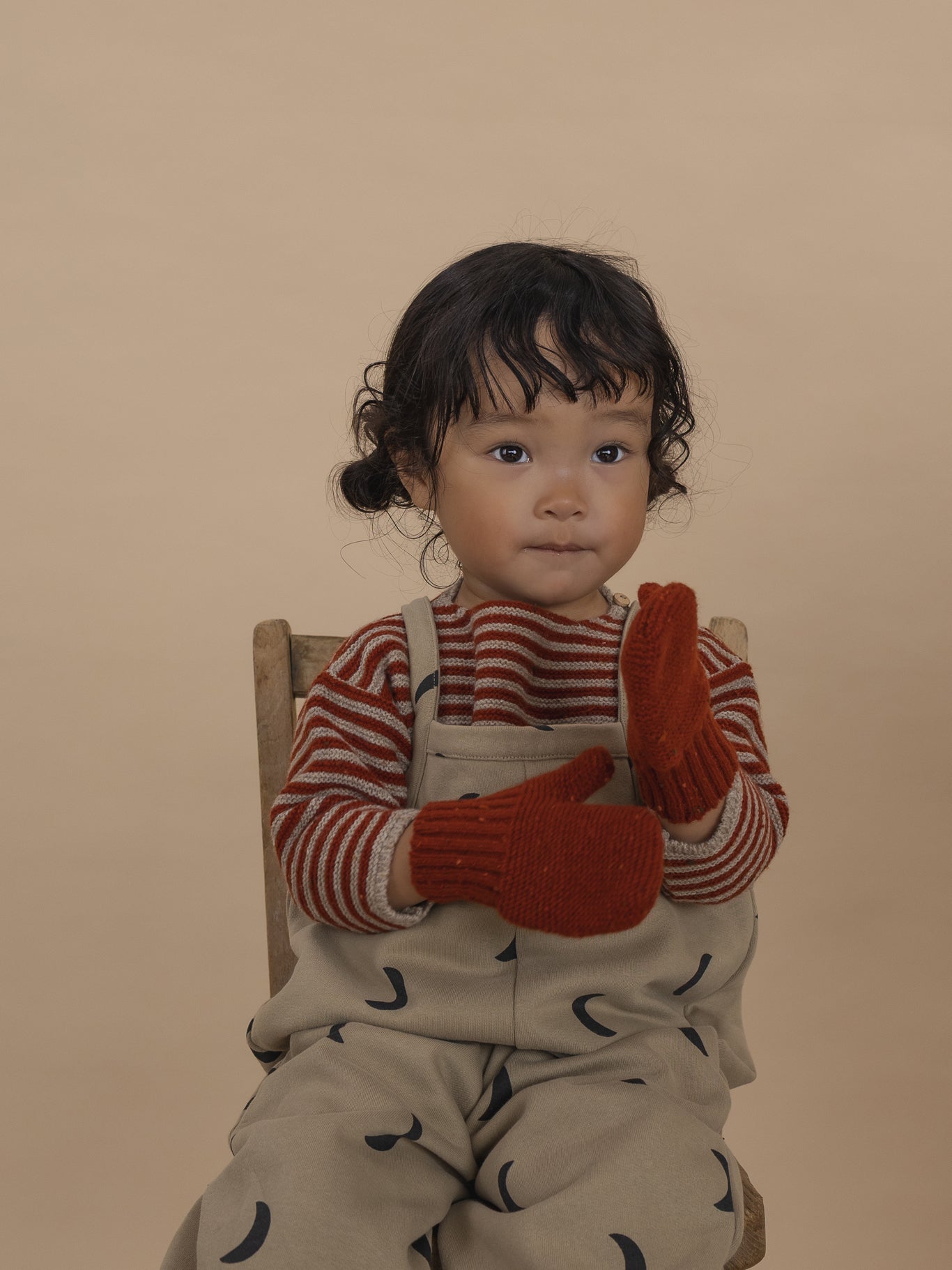 toddler in wool jumper with paprika red stripes, beige dungarees with moon print, and red mittens