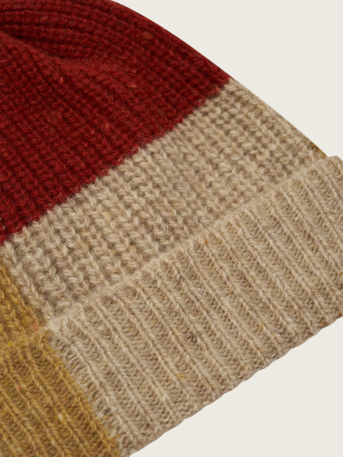 Close-up of a knitted beanie hat showing three color blocks: a dark red section at the top, a wide beige/tan section in the middle, and a mustard yellow section at the lower left, with a ribbed beige cuff on a white background.