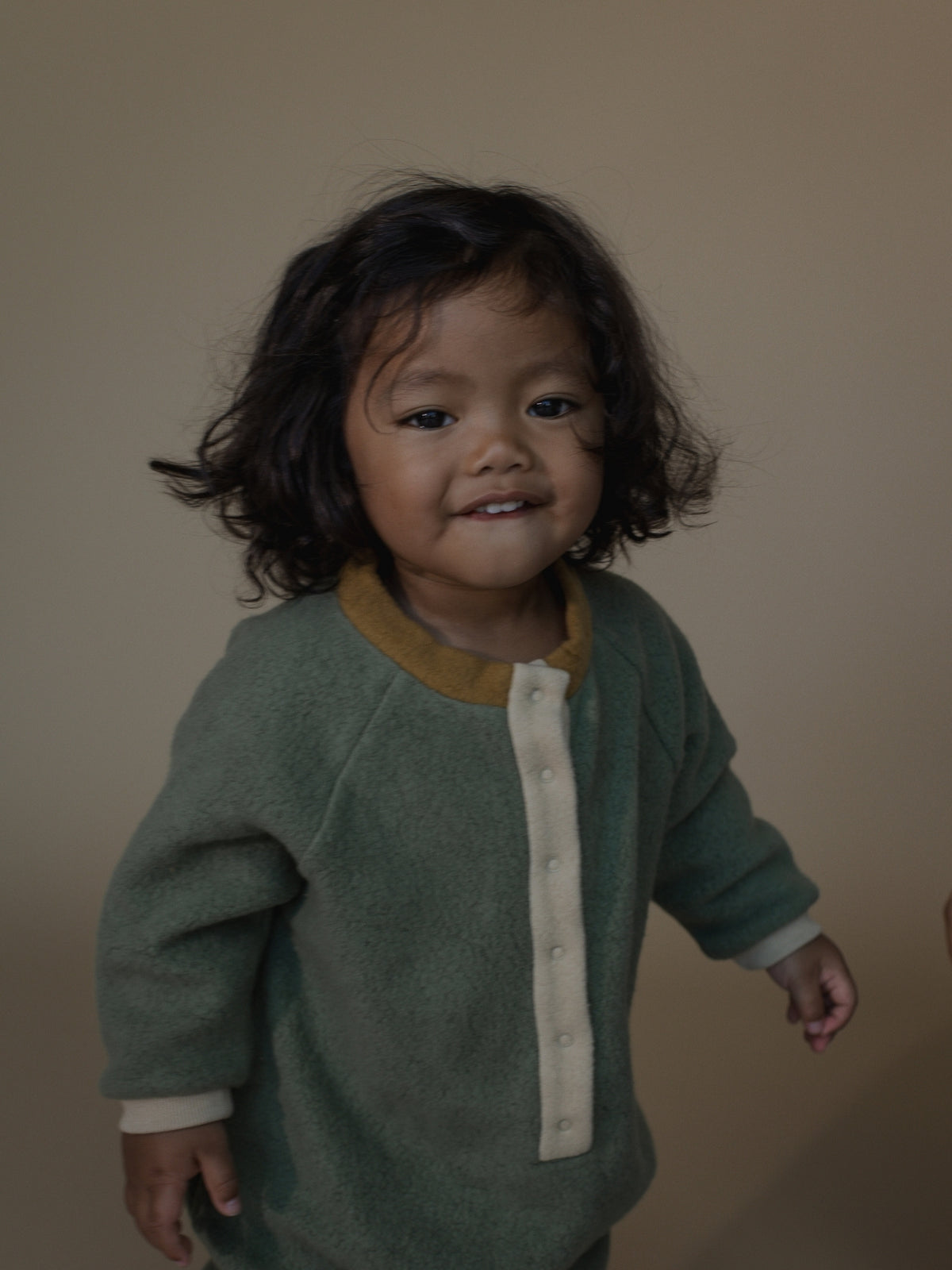 A smiling toddler with dark curly hair wearing a sage green knit cardigan with a mustard yellow collar and cream buttons, standing against a plain beige background.