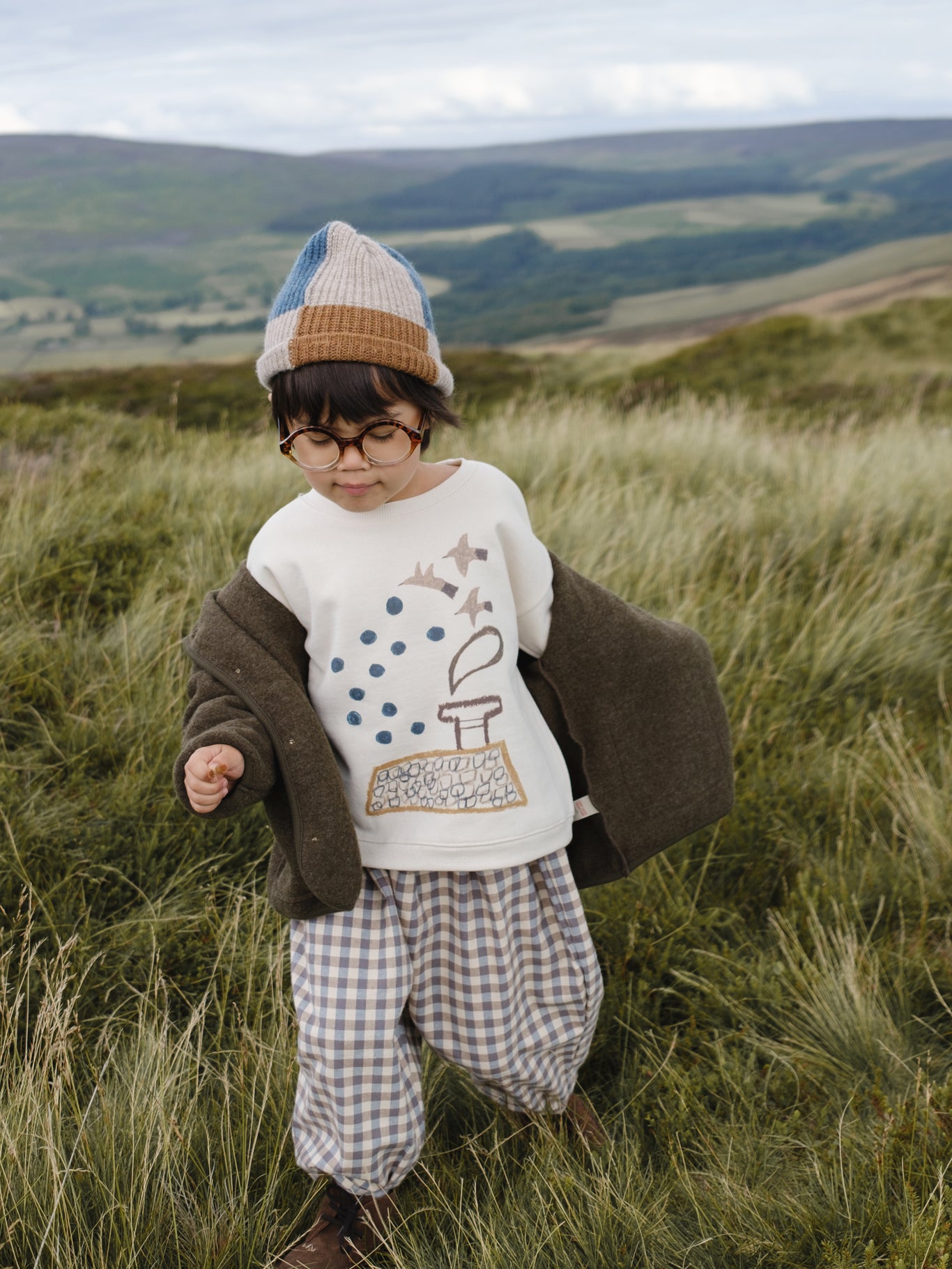 toddler in sweatshirt with bird graphic, gingham pants, patchwork beanie, and wool jacket in grassy meadow