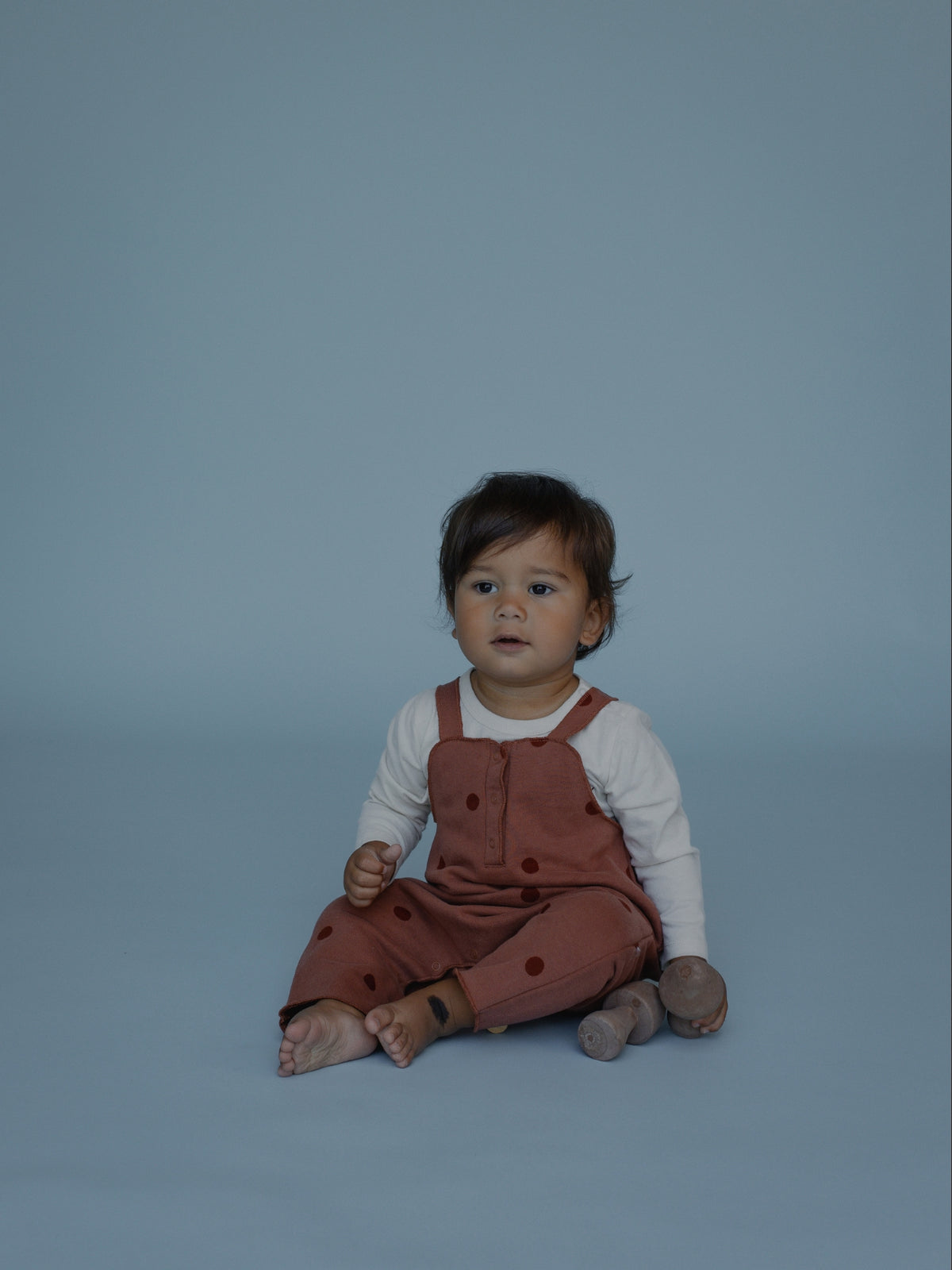 A baby sitting on a solid blue-gray studio background, wearing rust-brown polka-dot overall over a white long-sleeve shirt.