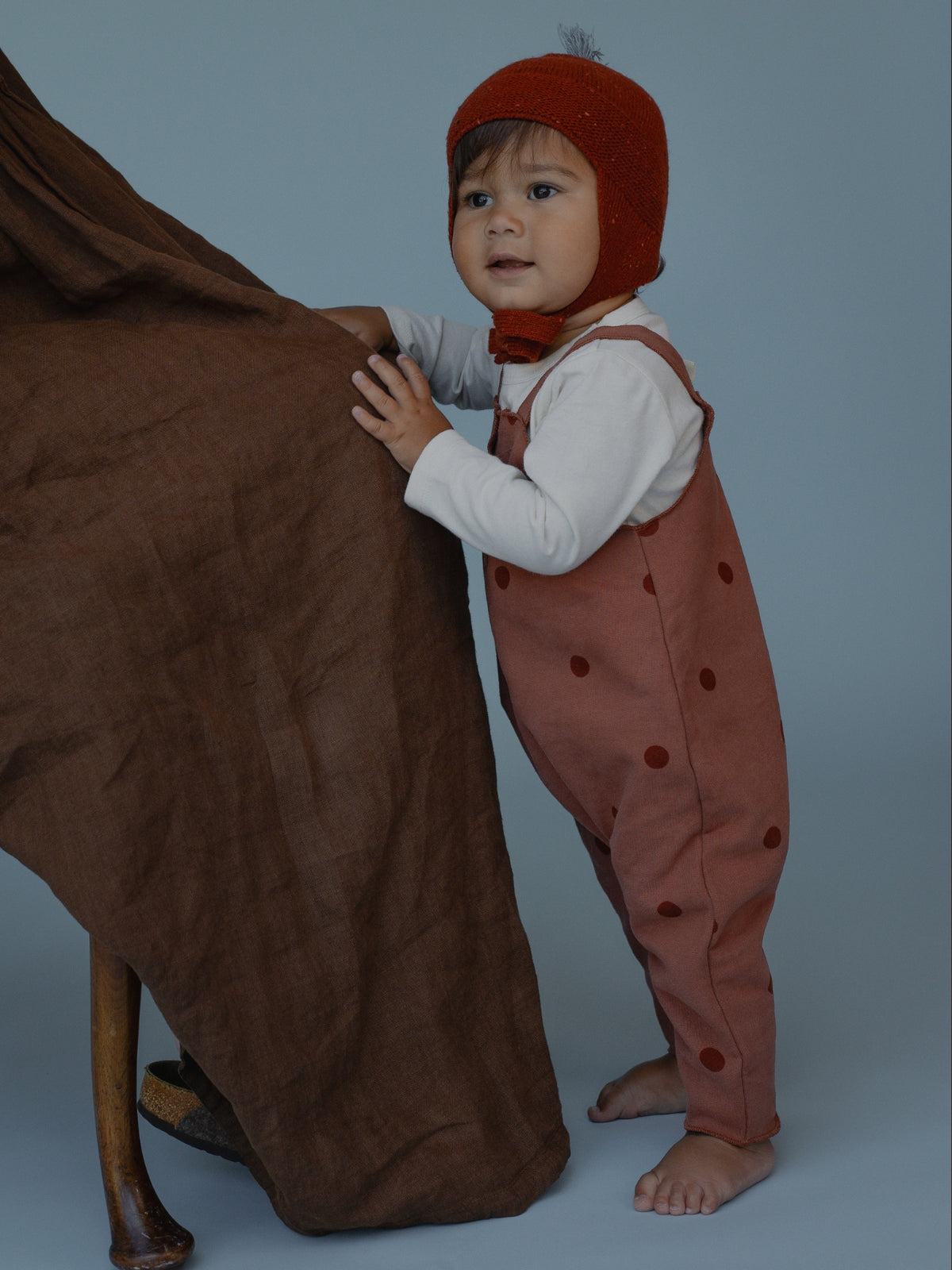 A baby wearing a rust-colored knit bonnet and matching polka-dot overalls over a white long-sleeve shirt, standing next to a large brown fabric drape on a blue-gray studio background