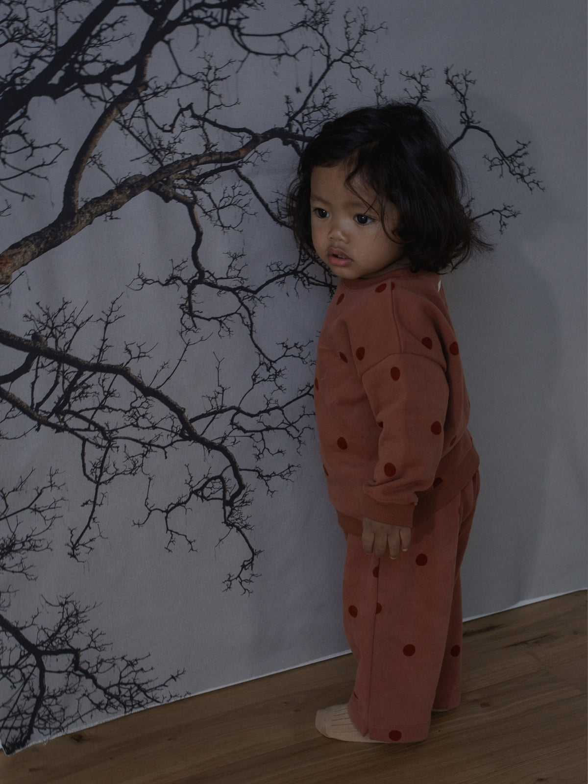 A toddler stands barefoot against a gray wall with a black tree branch print, wearing a rust-orange one-piece with dark red polka dots.