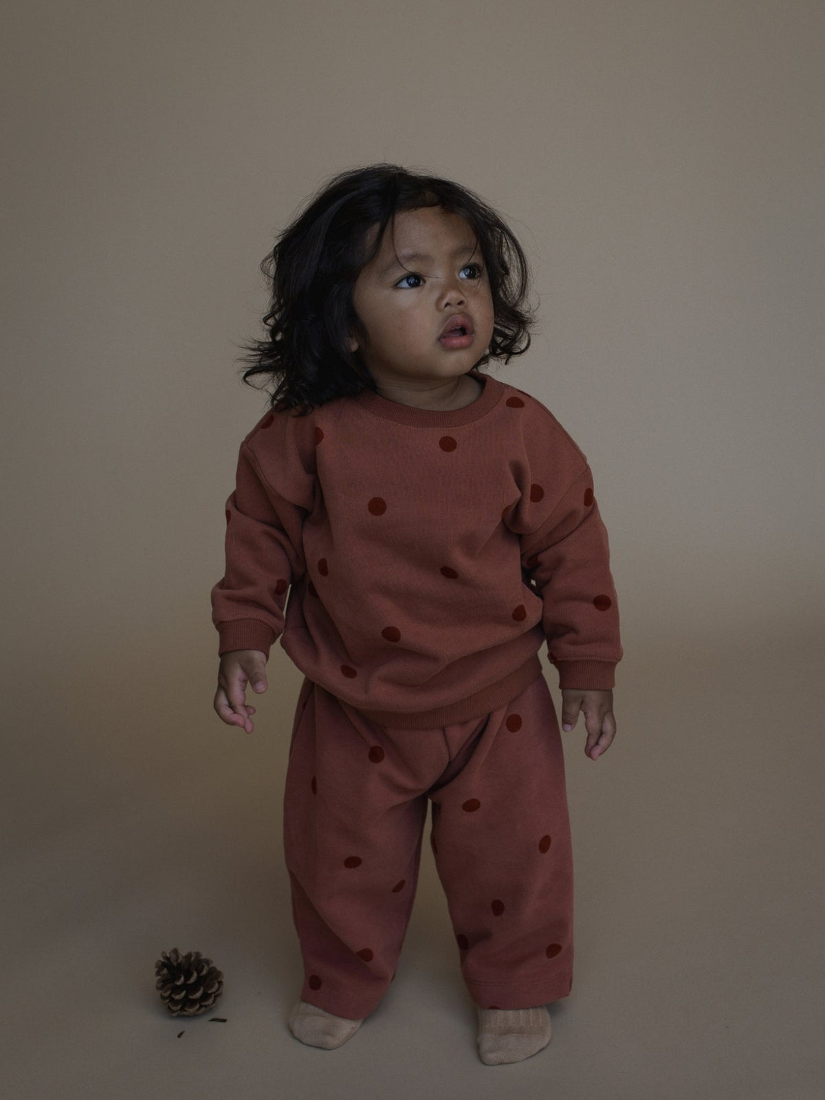 A toddler stands against a plain beige studio backdrop wearing a matching rust-brown long-sleeve top and pants set with darker brown polka dots. The child is barefoot, and a small pinecone sits on the floor nearby.