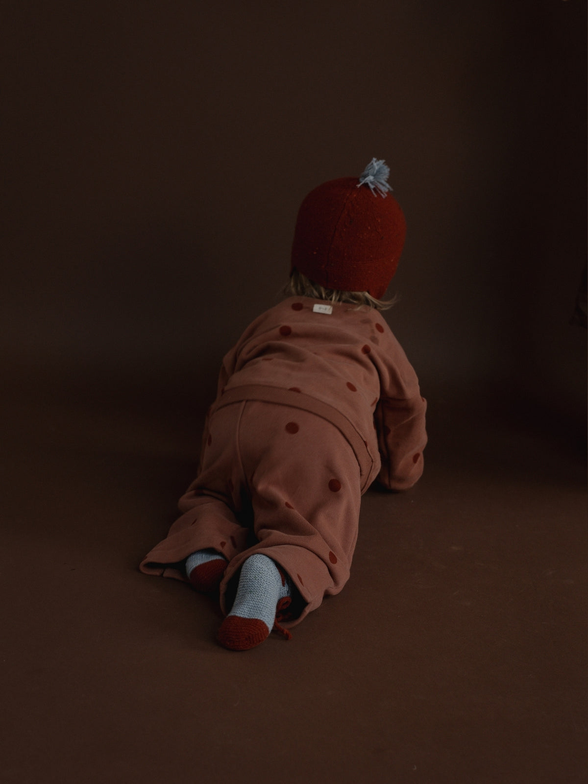 A baby seen from behind wearing a dusty rose polka dot romper, a red knit hat with a small blue bow, and light blue socks, crawling on a dark brown studio floor.