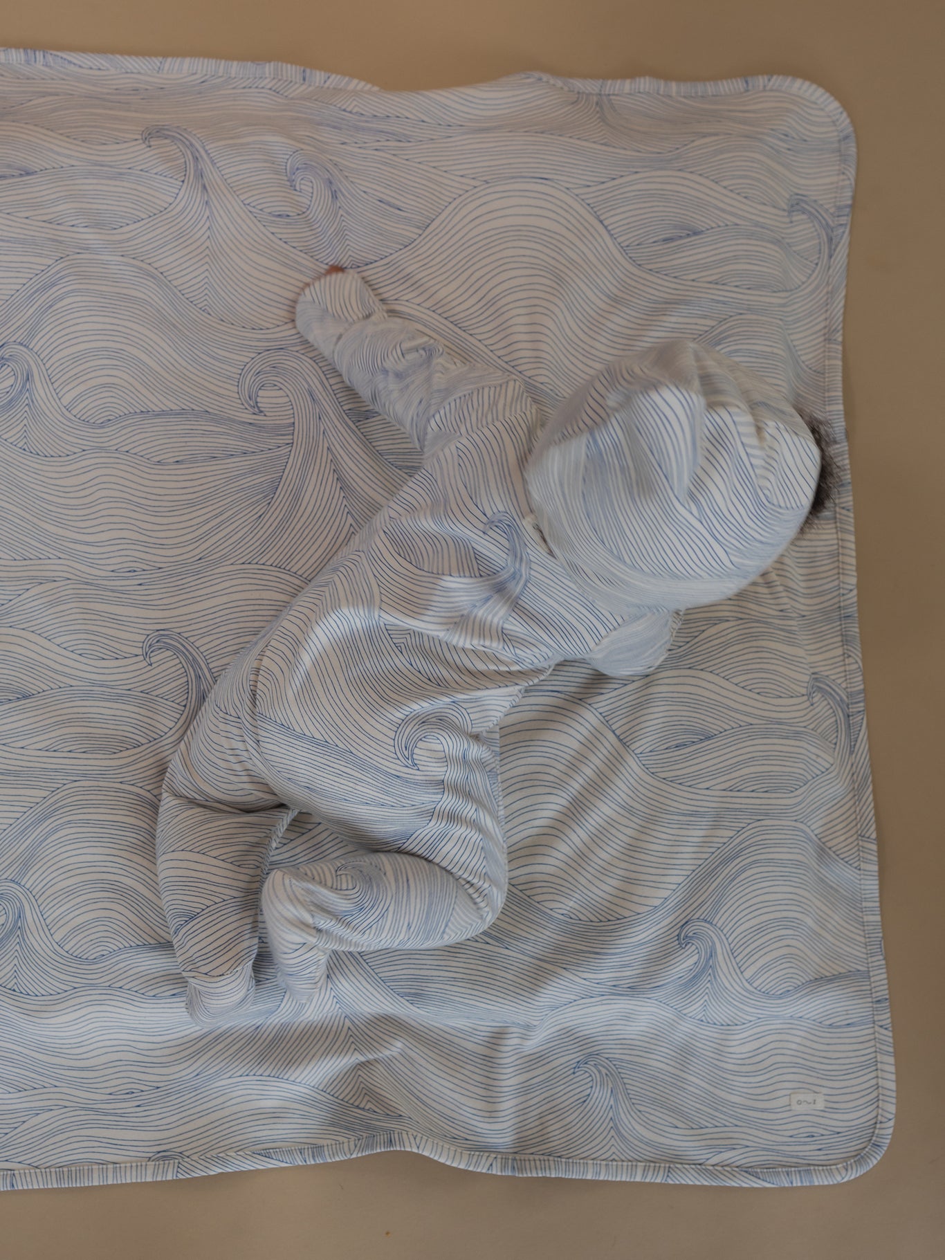 baby in cream pixie bonnet & playsuit with blue line-drawn ocean waves, lying on matching blanket