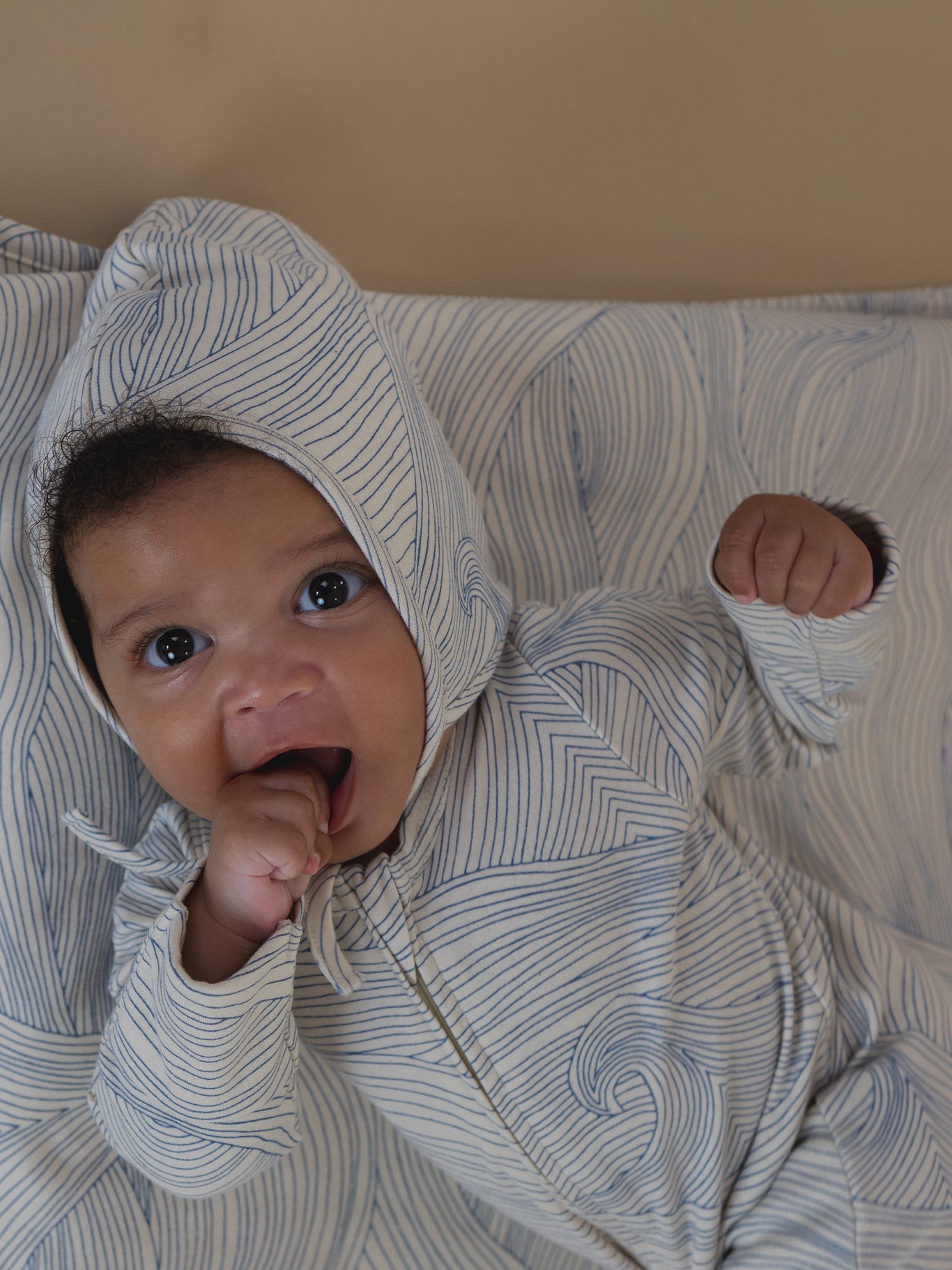 baby in matching cream undyed cotton pixie bonnet and zip playsuit with blue line-drawn ocean waves