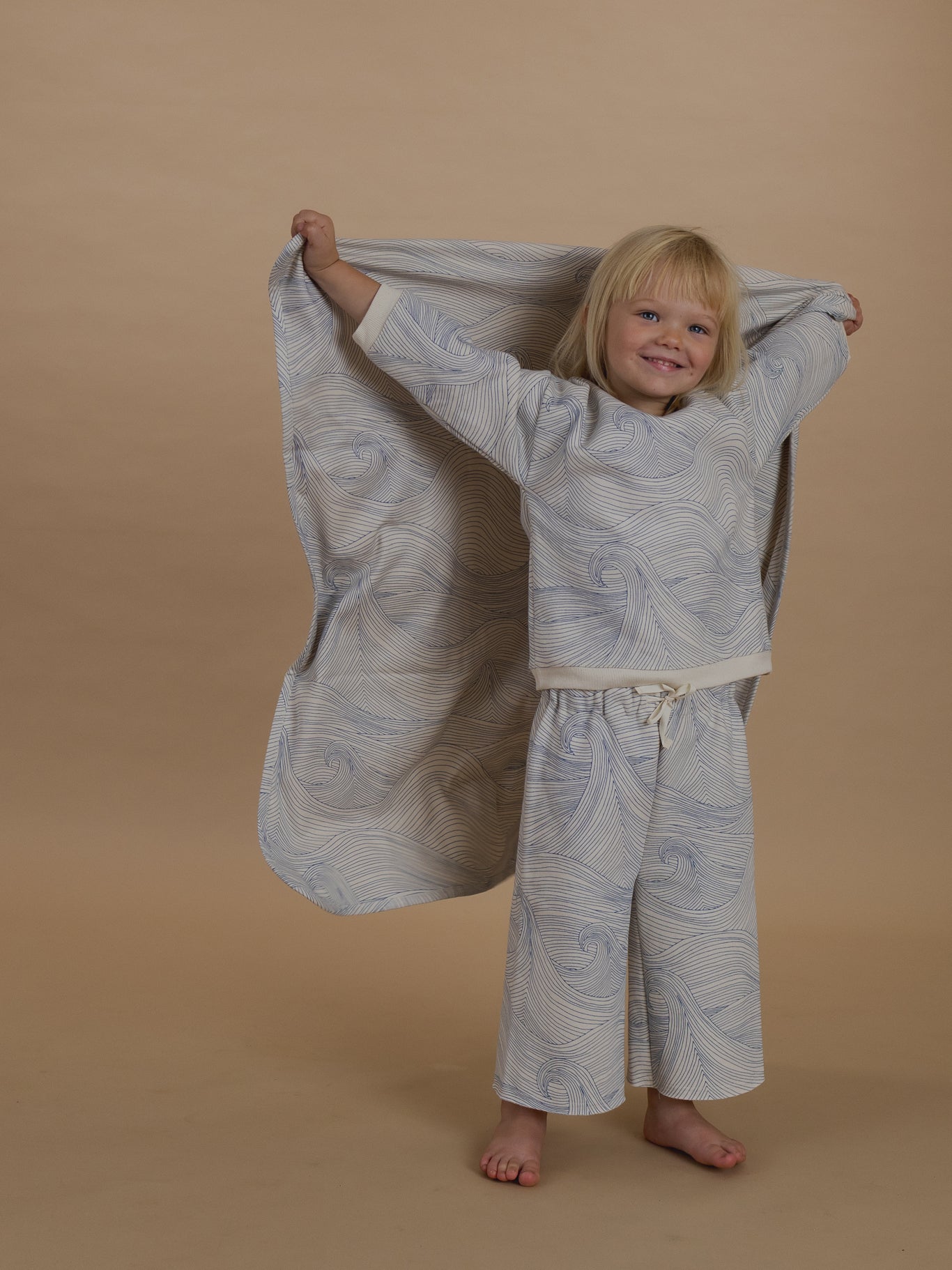 toddler holding cream blanket with blue line-drawn ocean waves on back, in matching sweatshirt and pants