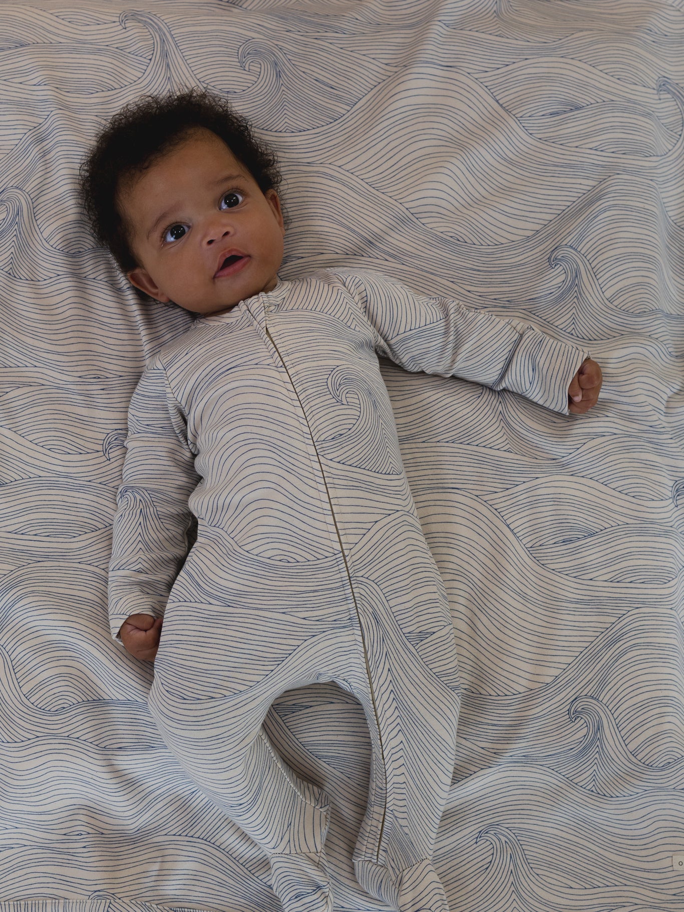 baby in undyed cotton zip sleepsuit with blue Seascape line-drawn ocean waves, and matching blanket