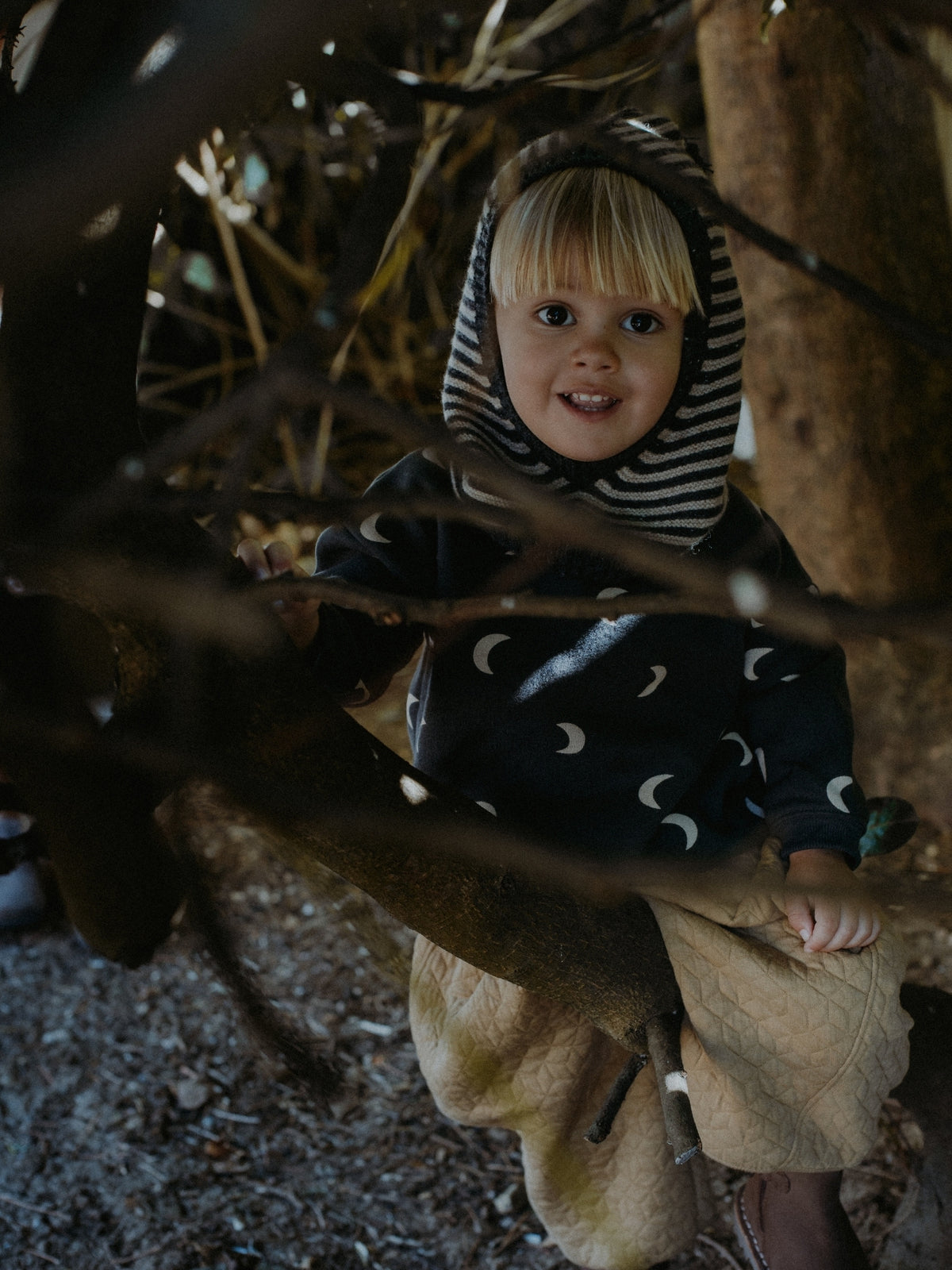 A young child wearing a cozy hooded outfit with a black and white striped hood and a dark blue moon-patterned top, sitting outdoors among tree trunks and branches.