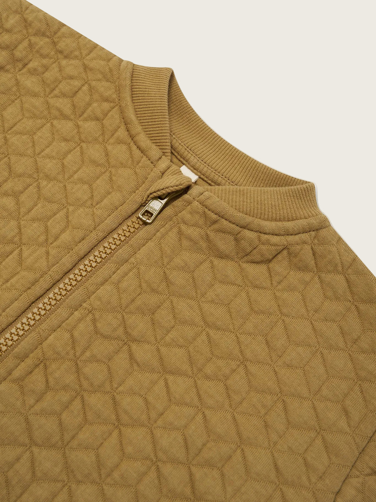 Close-up of a quilted zip-up jacket in a warm camel brown color, showing the geometric stitched pattern, ribbed collar, and a silver zipper pull on a white background.