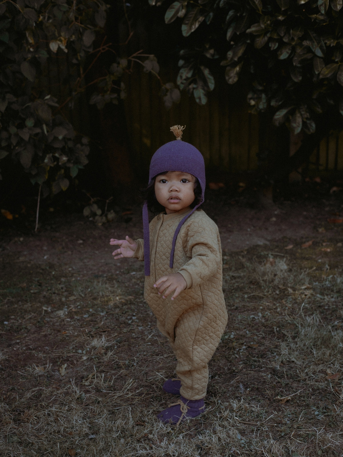 A toddler standing outdoors in a backyard wearing a tan quilted one-piece jumpsuit with purple suspenders and a matching purple knit hat with a small pom-pom.