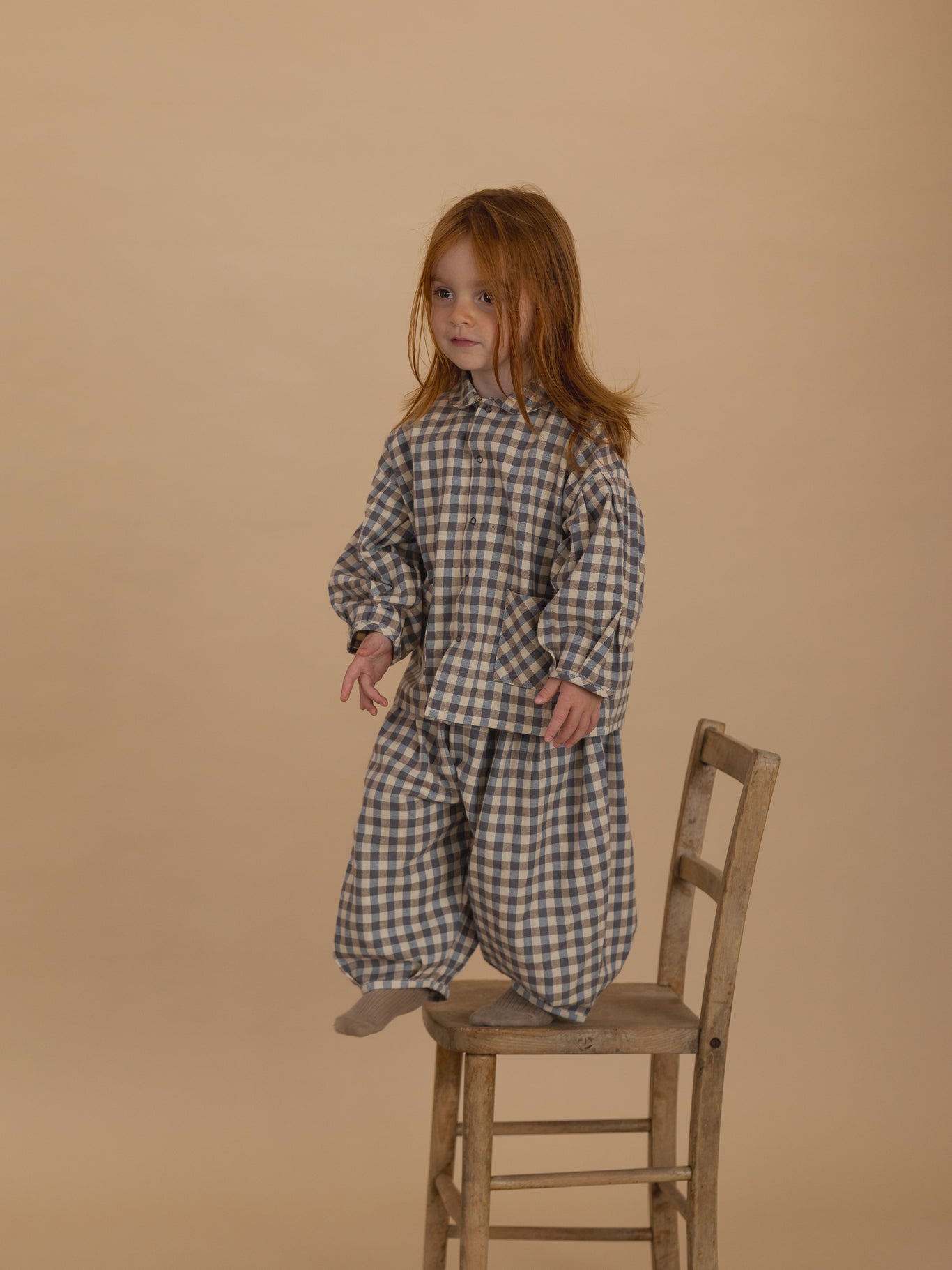 toddler in matching flannel balloon pants in cream and blue gingham and shirt, standing on chair