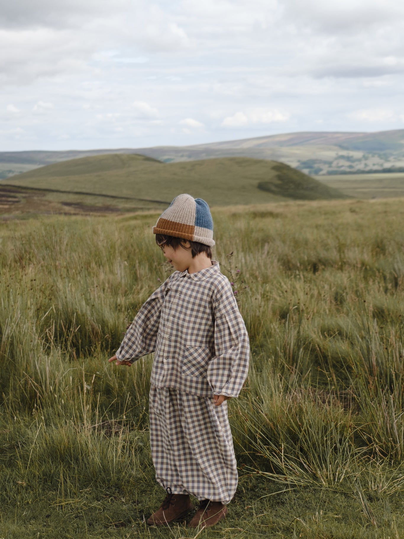 toddler in gingham lodge shirt with matching balloon pants & patchwork beanie, standing in grassy meadow
