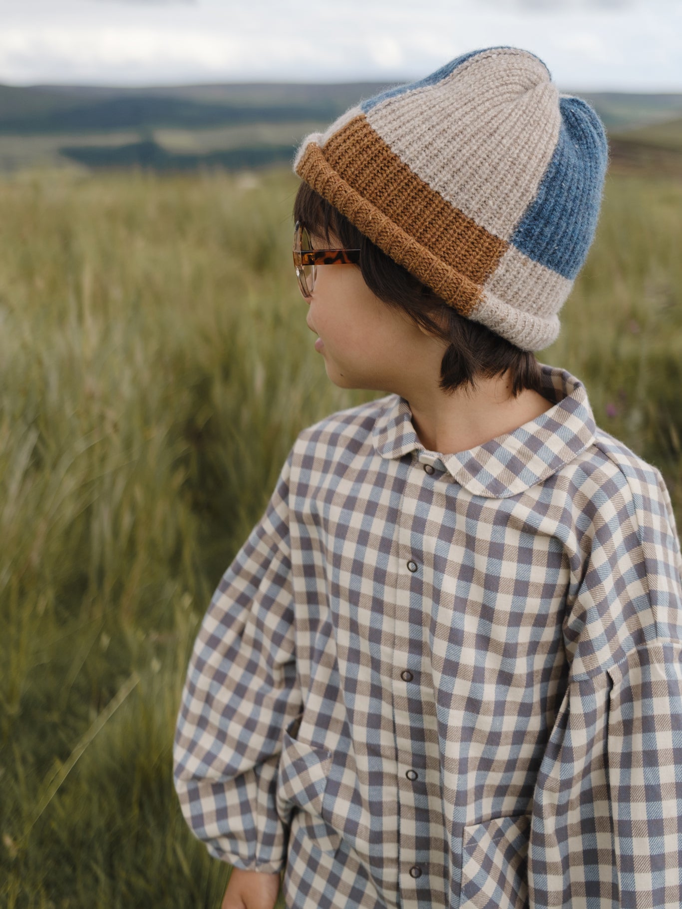 toddler in cream & blue gingham lodge shirt, patchwork beanie & glasses standing in grassy meadow