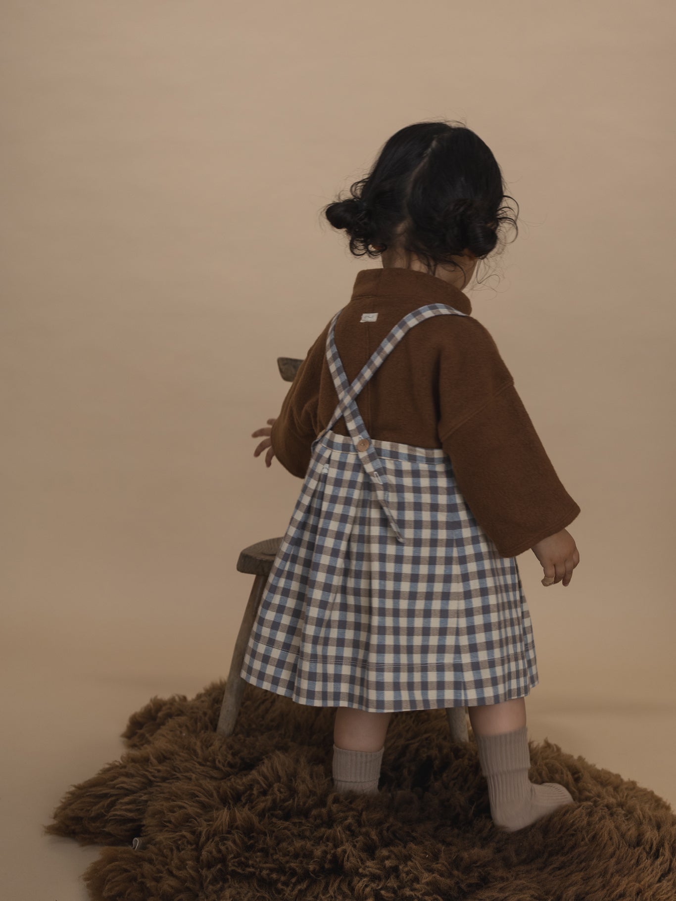 back of a toddler in cream and blue gingham pleated pinafore skirt with adjustable straps, and jumper