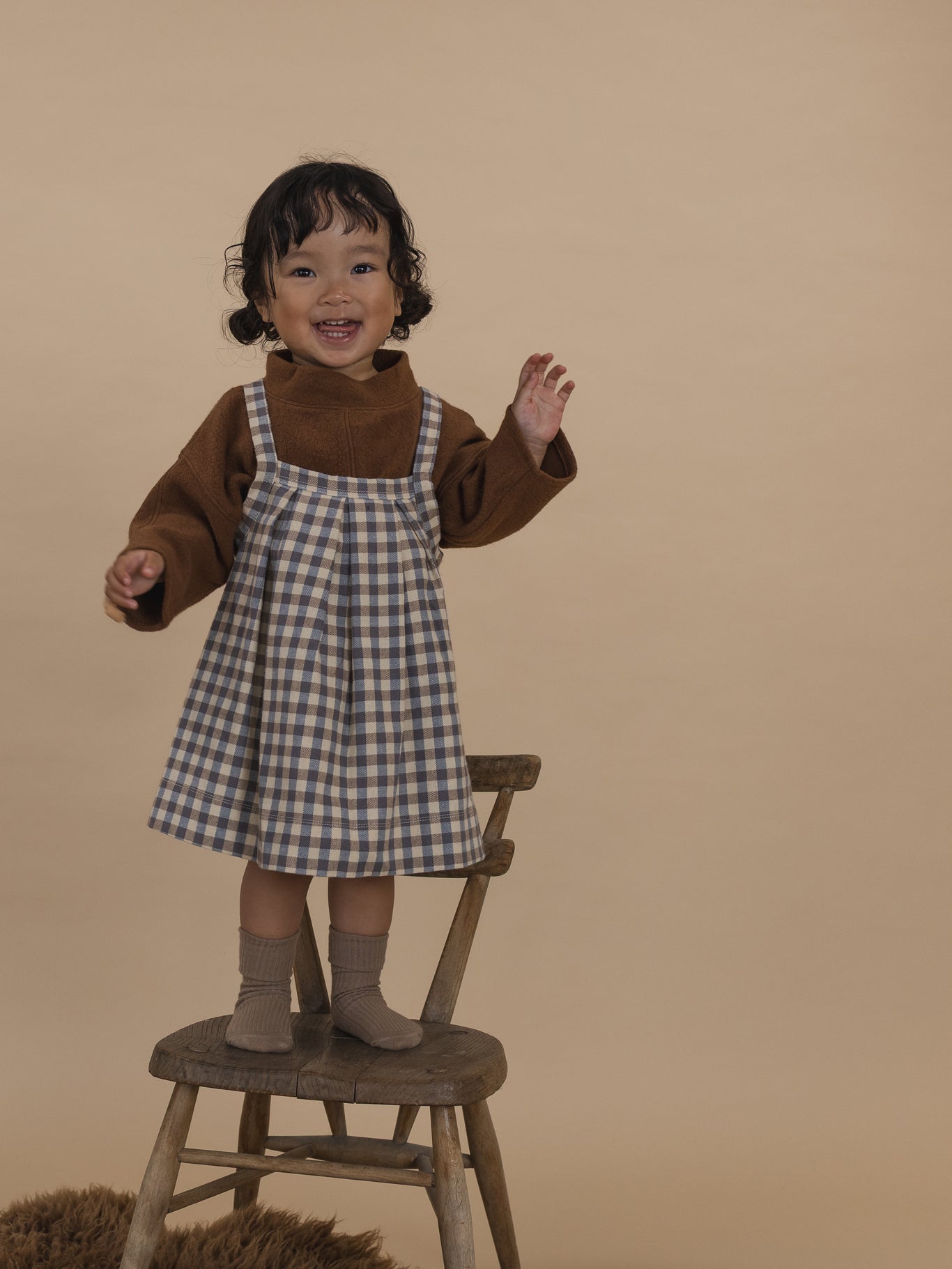 toddler in cream and blue gingham pleated pinafore skirt with brown jumper, standing on wooden chair