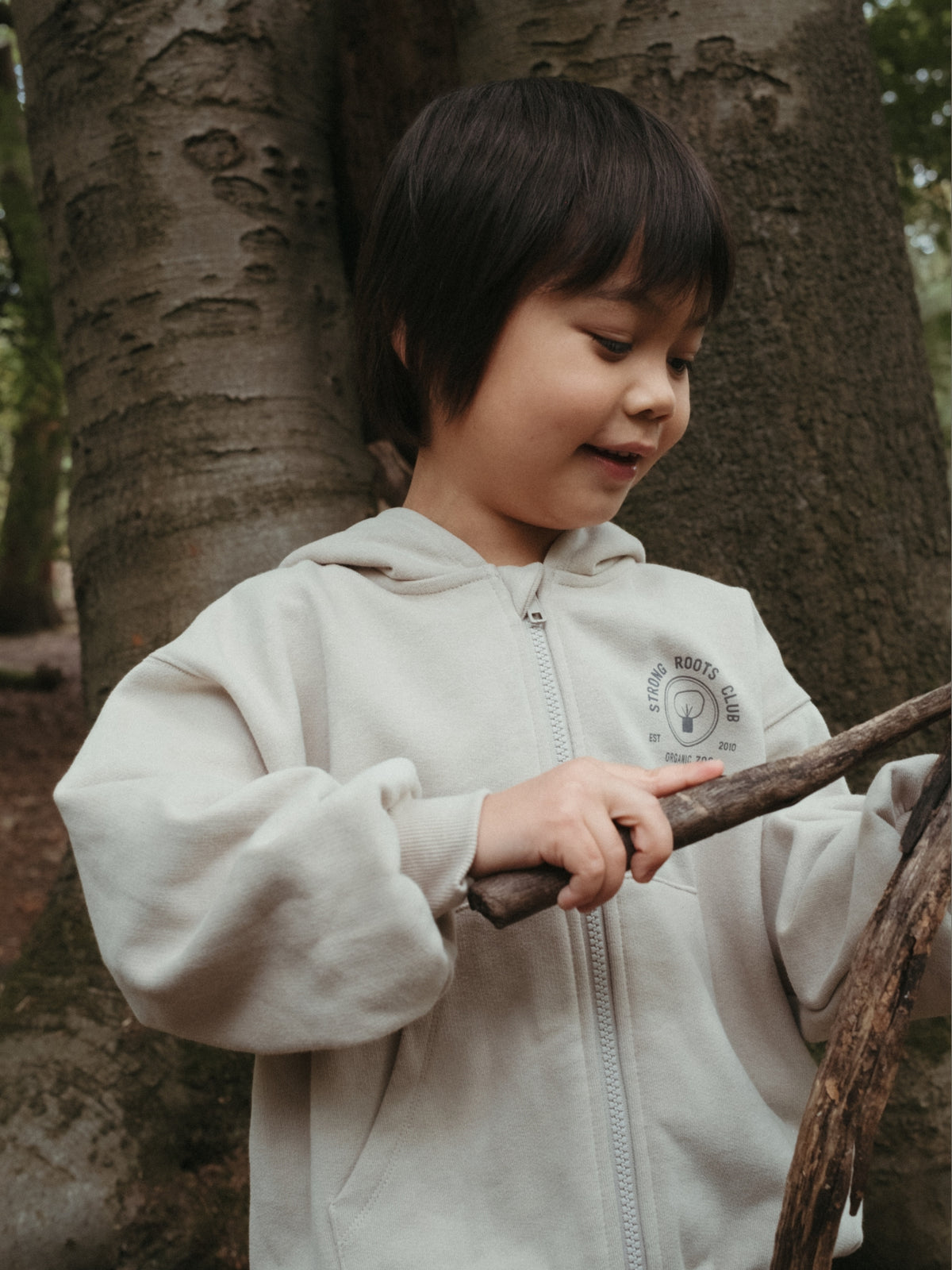 A young child wearing a cream-colored zip-up hoodie stands outdoors in front of a large tree trunk, holding a piece of wood.