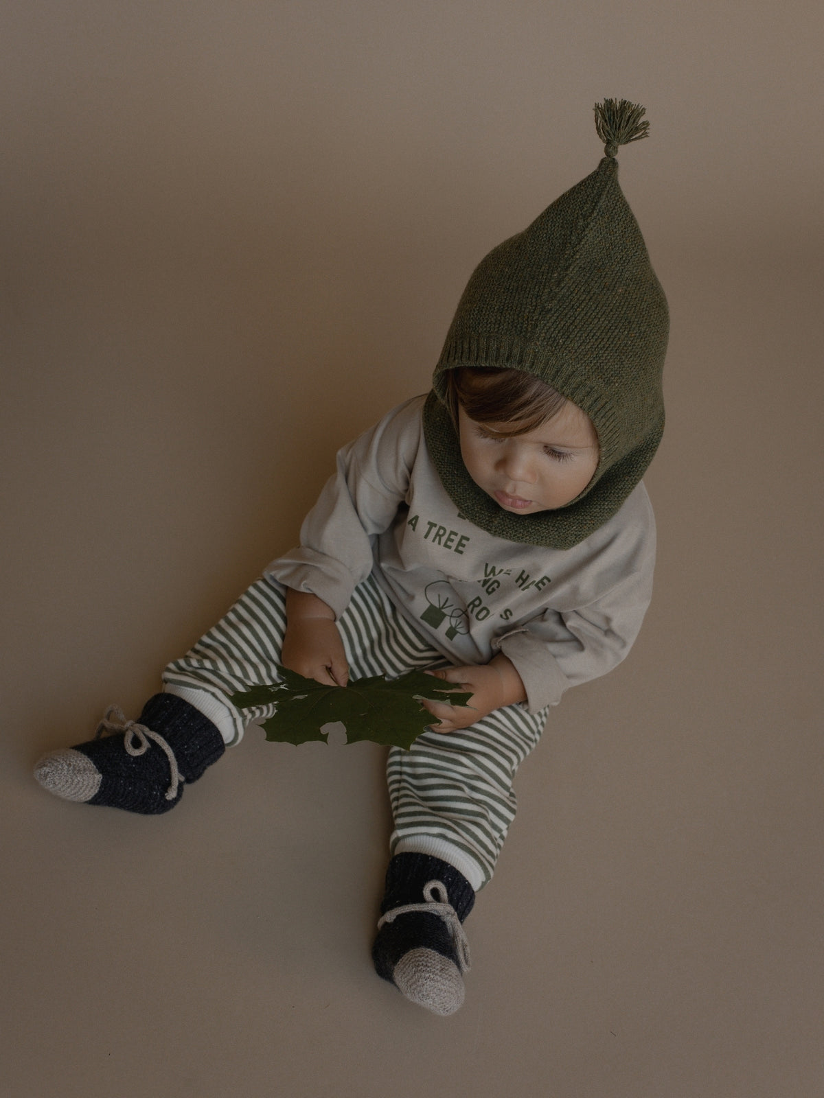 A baby wearing a dark olive green knit bonnet with a pom-pom and a matching knit neck warmer, paired with a light gray long-sleeve shirt, green and white striped pants, and navy socks, sitting on a solid beige background.