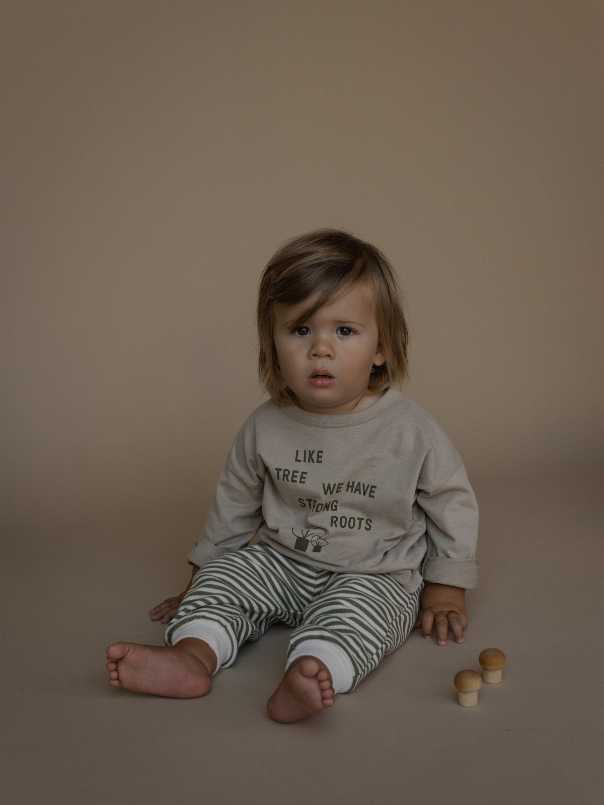 A toddler sitting on a warm beige studio floor wearing a light beige long-sleeve shirt with the text 'LIKE TREE WE HAVE STONED ROOTS' and green-and-white striped pants. The child is posed with small wooden toy mushrooms beside them against a solid beige backdrop.
