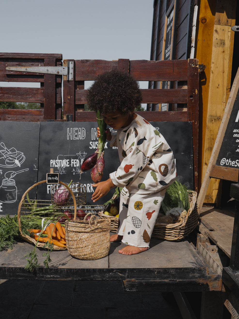 toddler in wide leg pants & sweatshirt with farmers market print taking shallots from basket
