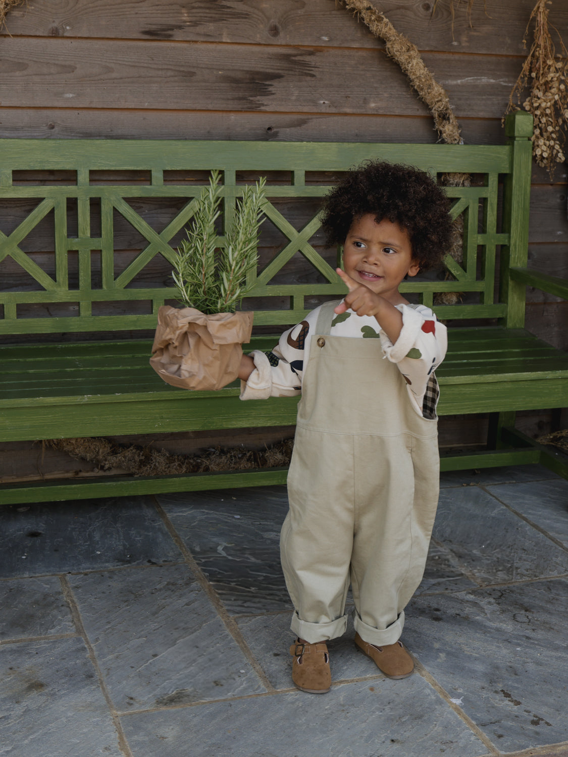 toddler in oversized grey cotton twill dungarees & sweatshirt with prints holding paper bag with herbs