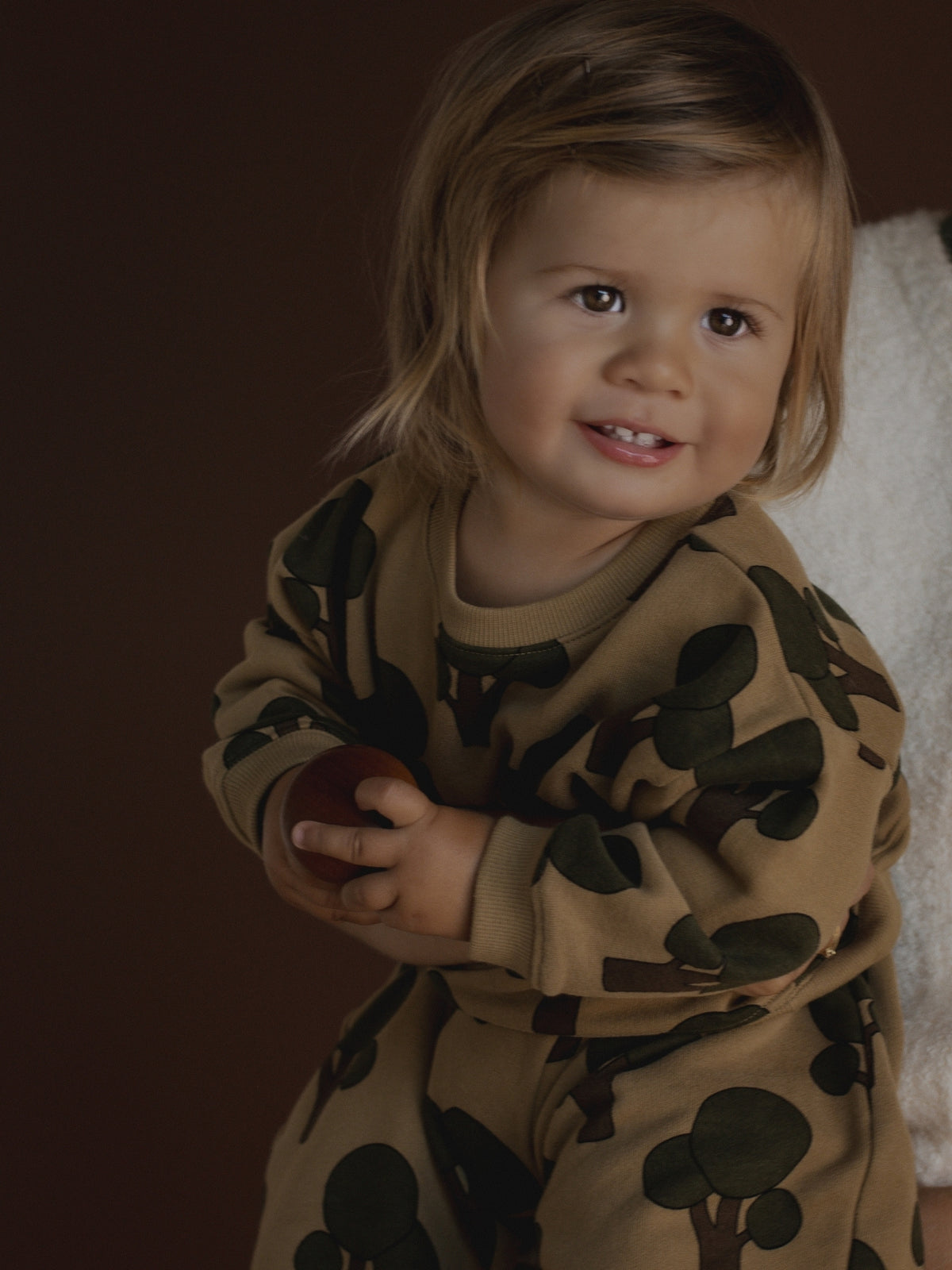 A toddler wearing a tan and dark green tree-print set, holding a small red object against a dark brown studio background.