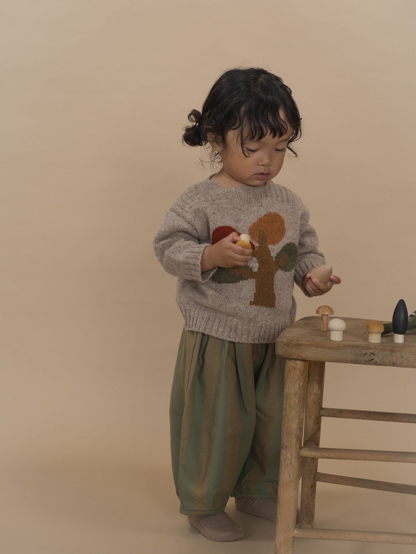 toddler in beige wool jumper with tree motif and balloon pants, playing with wooden toys
