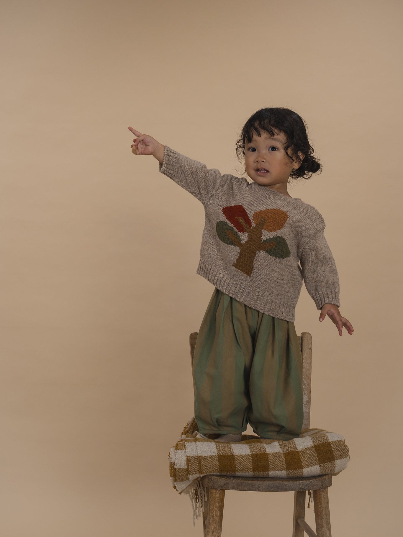 toddler in beige wool jumper with tree motif and balloon pants, standing on chair and pointing