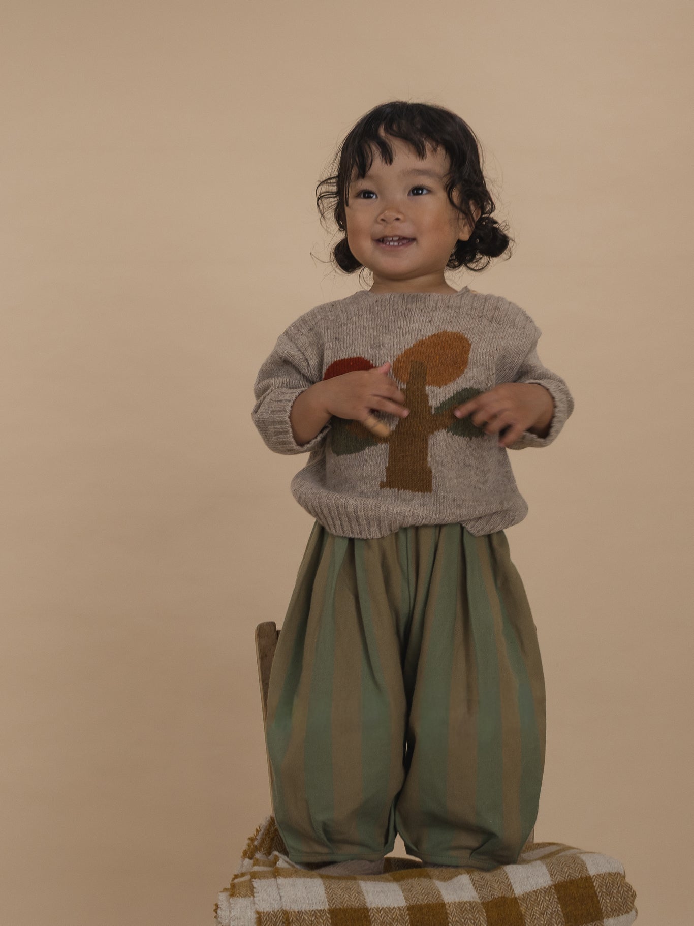 toddler in beige wool jumper with tree motif and striped balloon pants, standing on chair