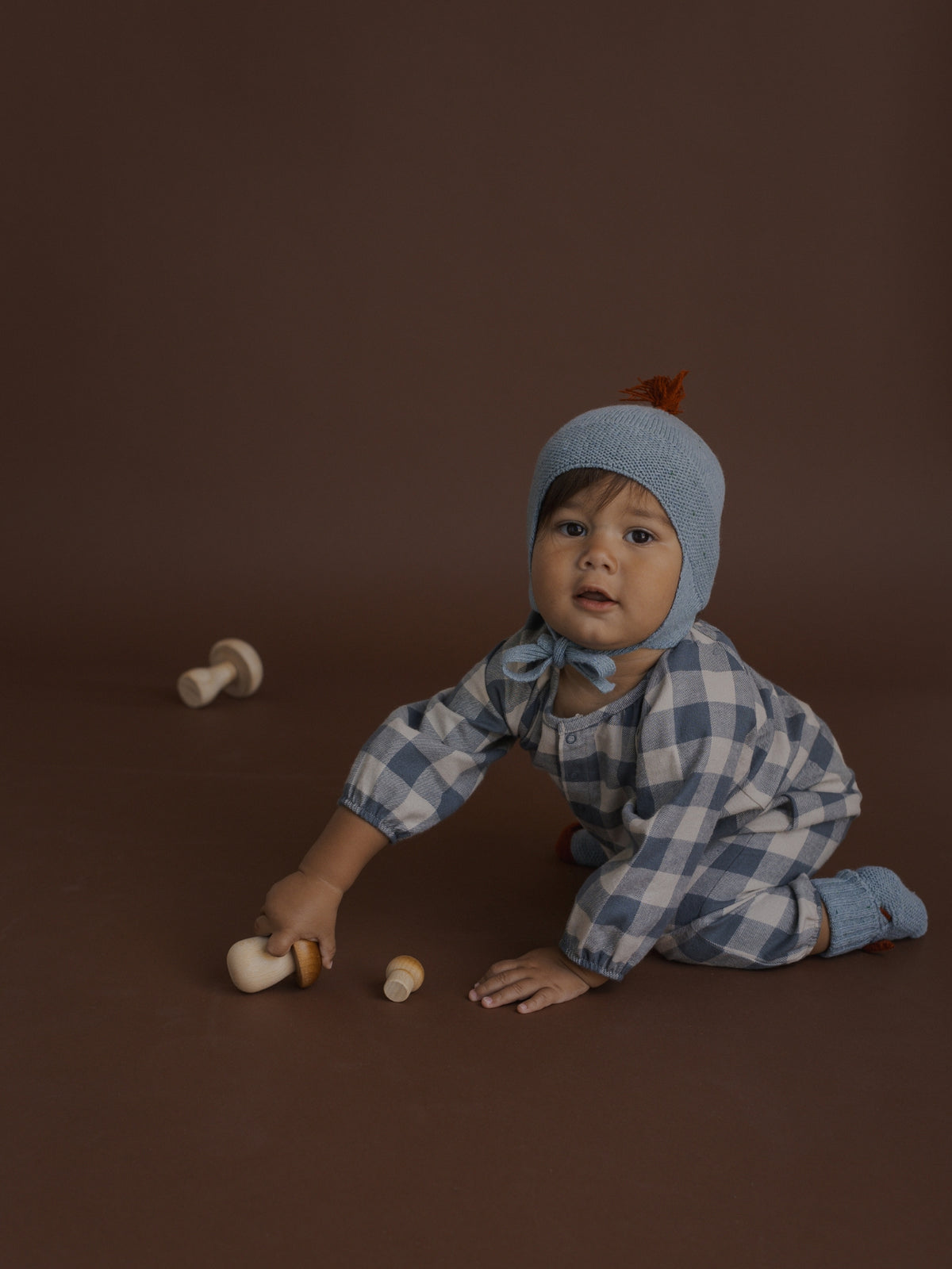 A baby wearing a light blue knit bonnet and a matching blue and white checkered romper, kneeling on a dark brown studio floor while touching small wooden toy mushrooms.