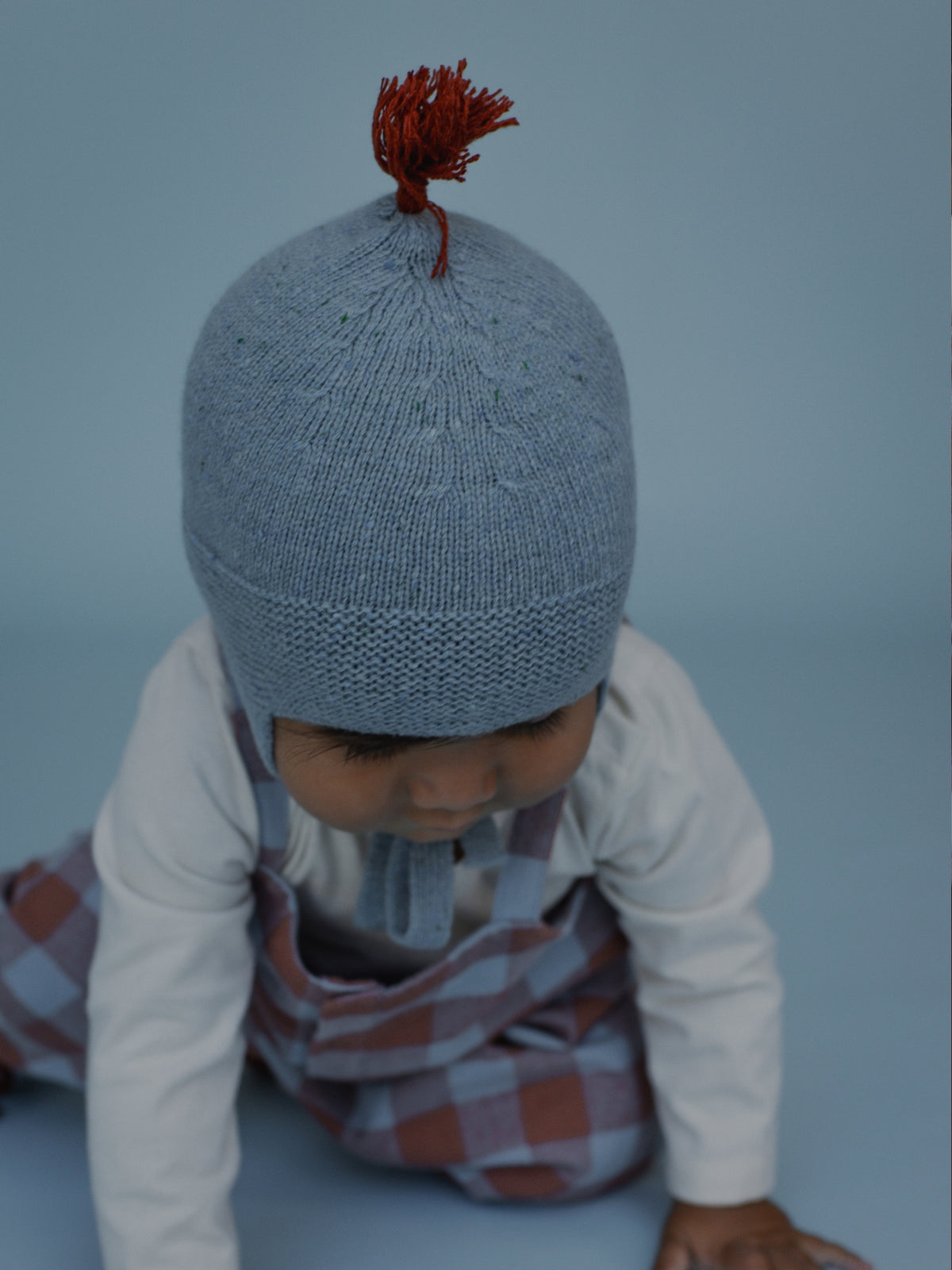 A baby wearing a light blue-gray knit beanie with a small rust-red tassel on top, crawling on a light blue studio floor.