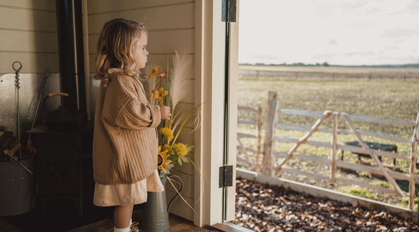 Child in almond corduroy dress with light brown quilt cardigan standing by a door looking at a field