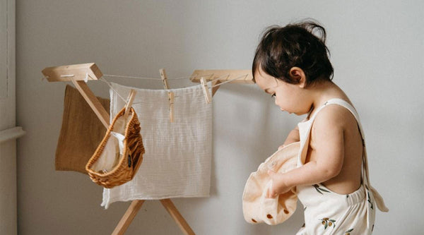 Toddler wearing sleeveless bodysuit holding a bucket hat standing next to laundry rack with muslin cloths