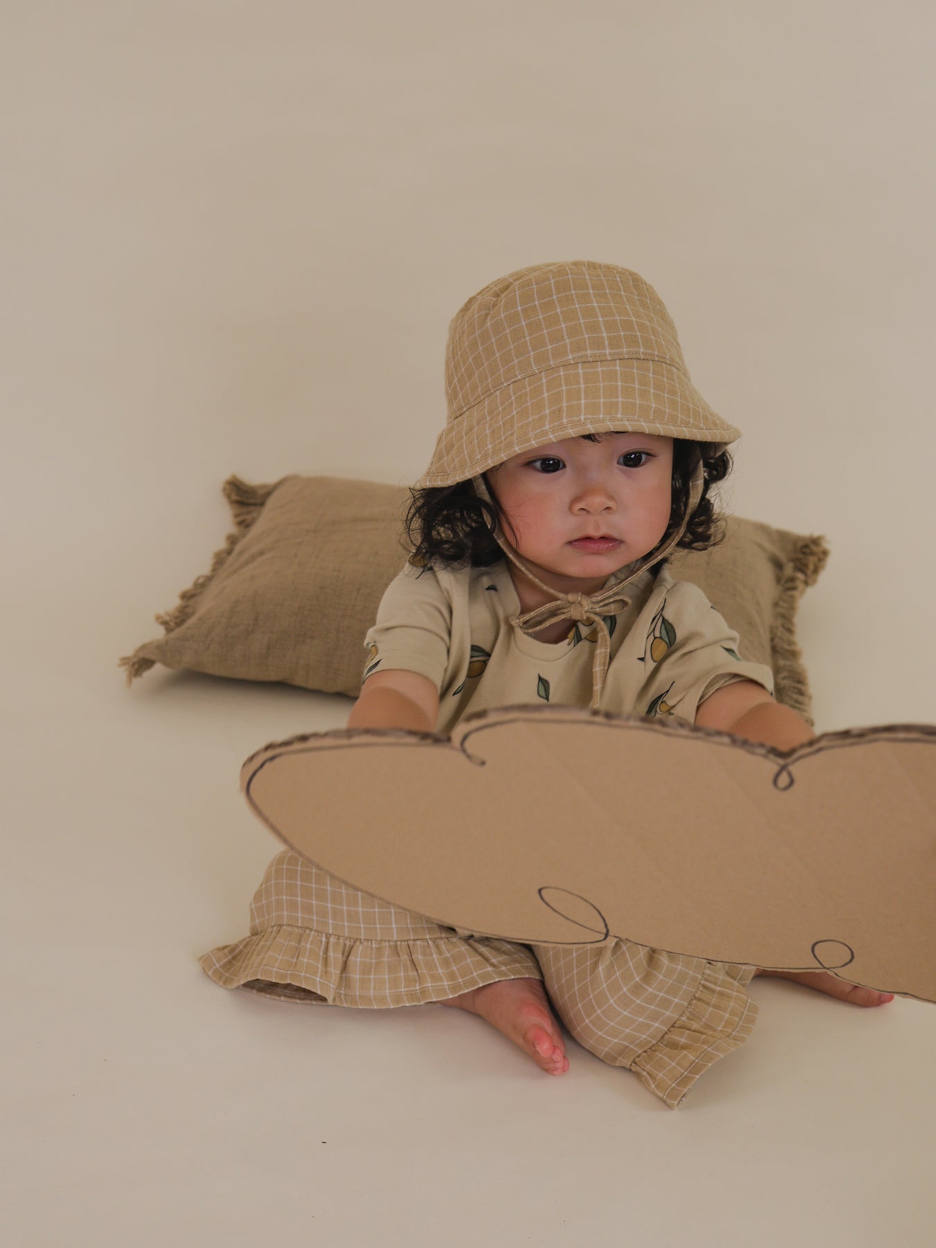toddler wearing wheat gold grid check bucket hat with straps tied under chin, sitting on floor holding cardbox cutout