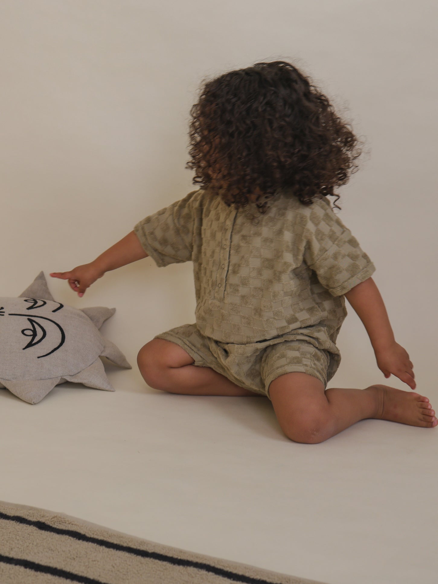 toddler in beige organic terry romper with checkerboard pattern & front poppers pointing to cushion