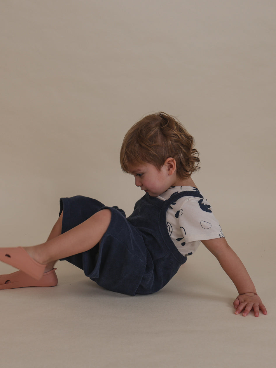 toddler in navy terry cropped overalls, t-shirt & swimming fins sitting on the floor leaning on hands