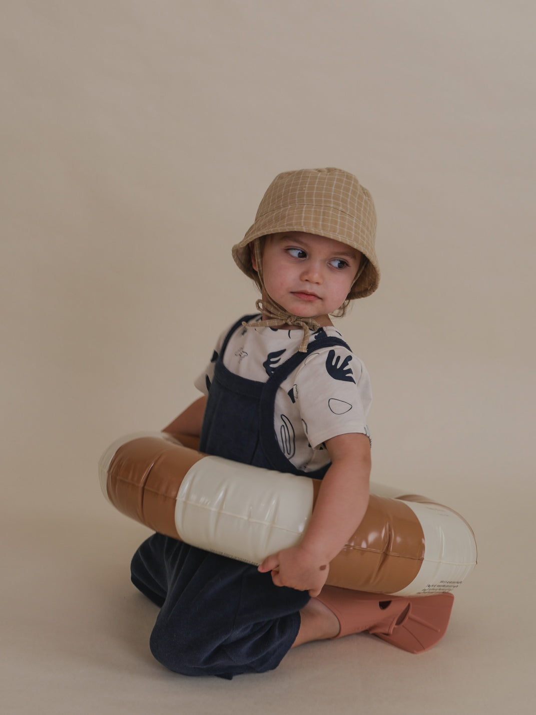 toddler in navy terry cropped dungarees, t-shirt, hat & flippers, holding pool ring at waist kneeling on the floor